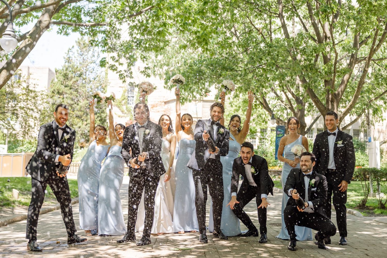 Wedding party celebrates with confetti throwing under green trees.