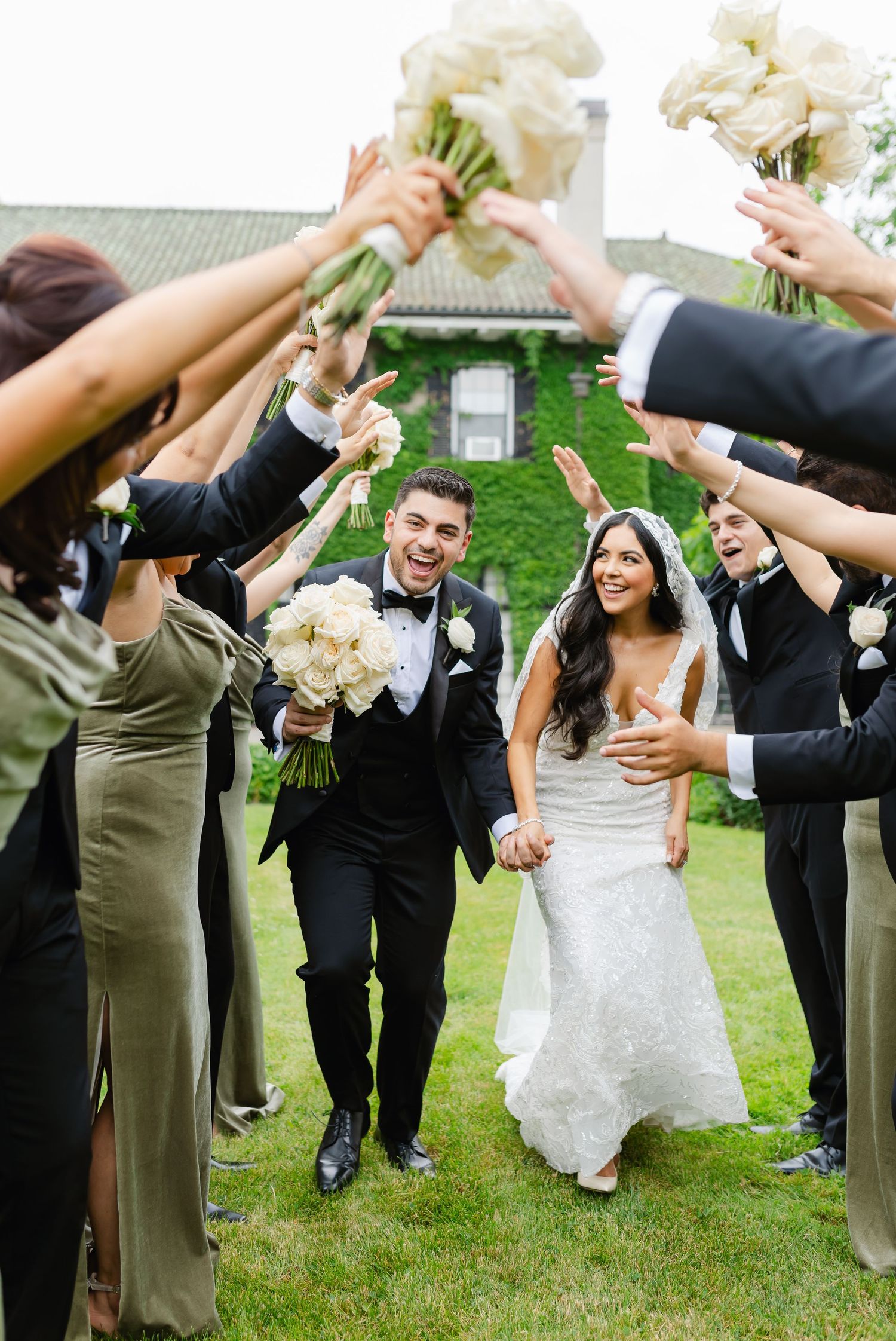 Wedding party lifts floral bouquets in celebration while walking on grass outside a historic venue.