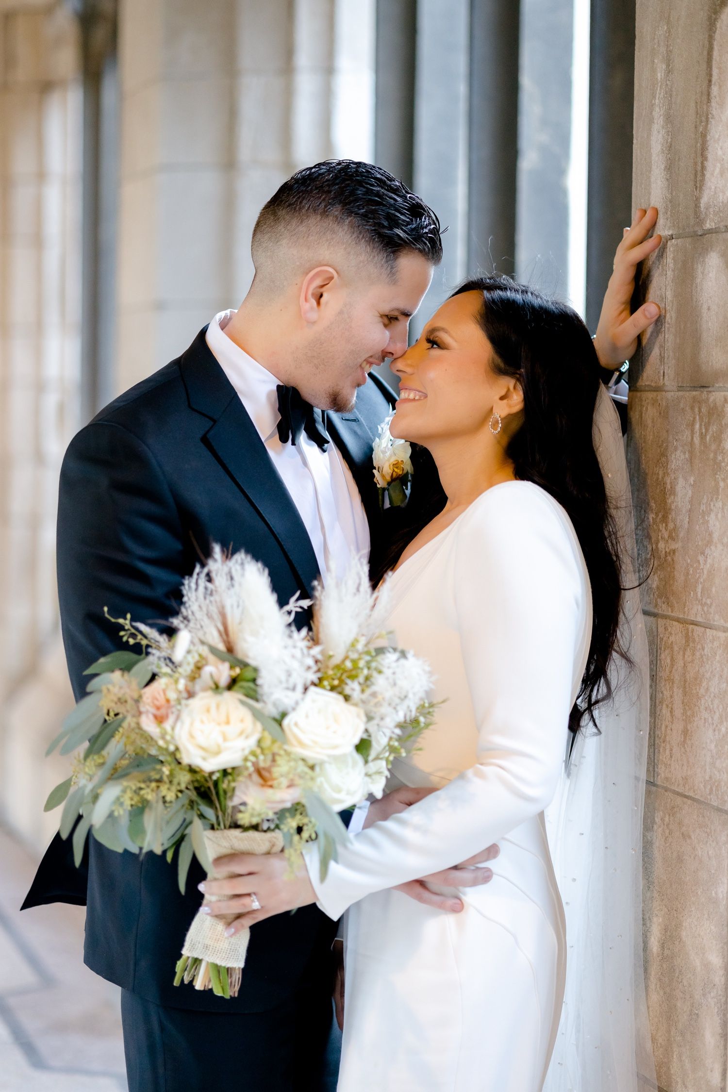 A joyful wedding portrait captured against stone architecture with a white and green bouquet.