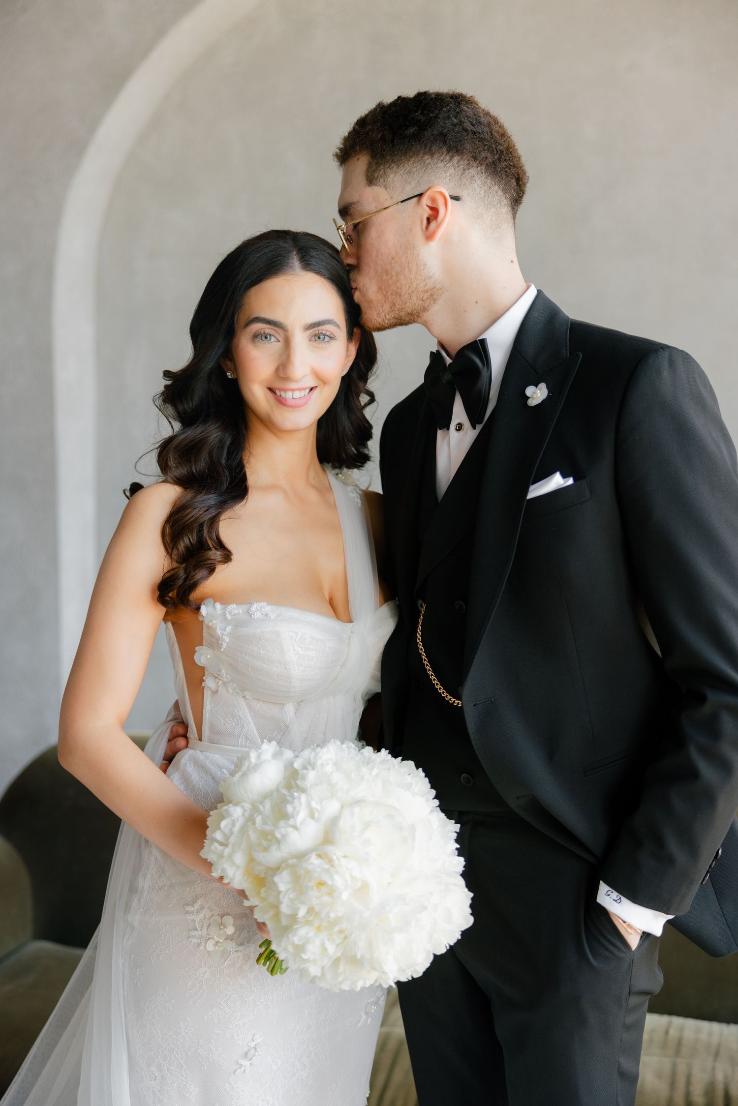 Romantic wedding portrait with bride holding white bouquet against arched wall.