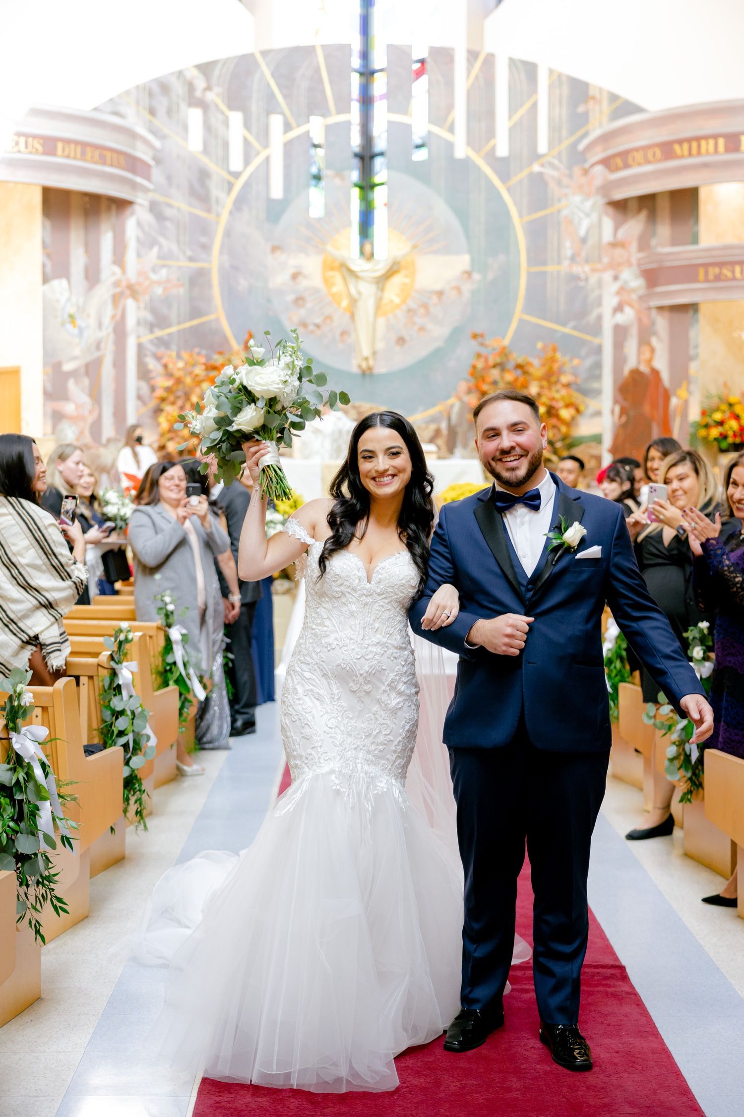 Newlyweds walk down red carpet aisle decorated with floral arrangements in ornate church setting.