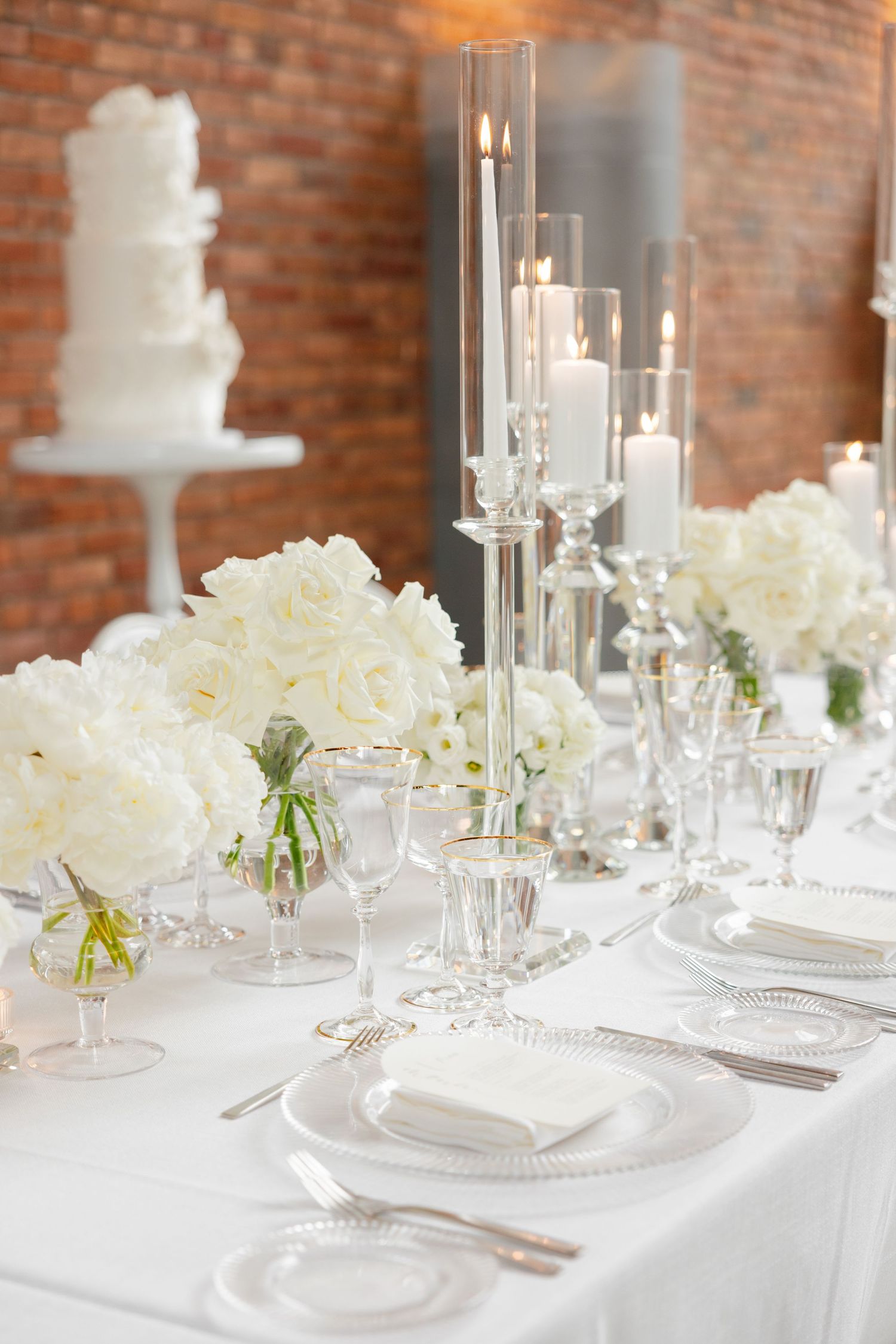 Wedding table setting with glass taper candle holders, white flowers, and exposed brick backdrop.