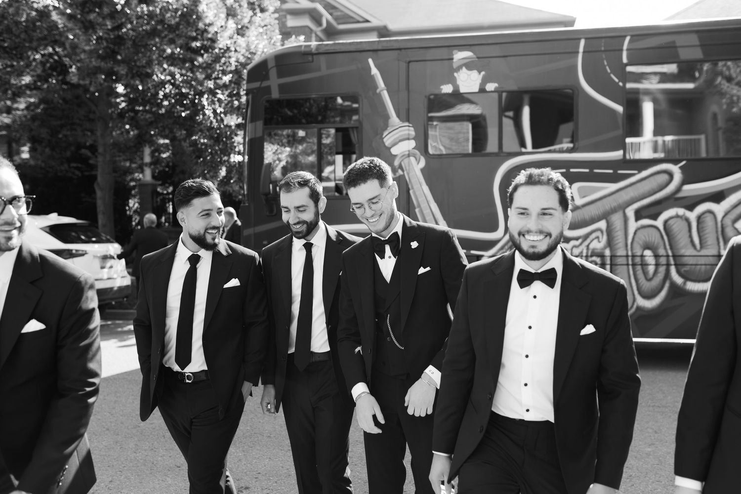 Wedding party members in formal attire gather beside a large vehicle in black and white photography.