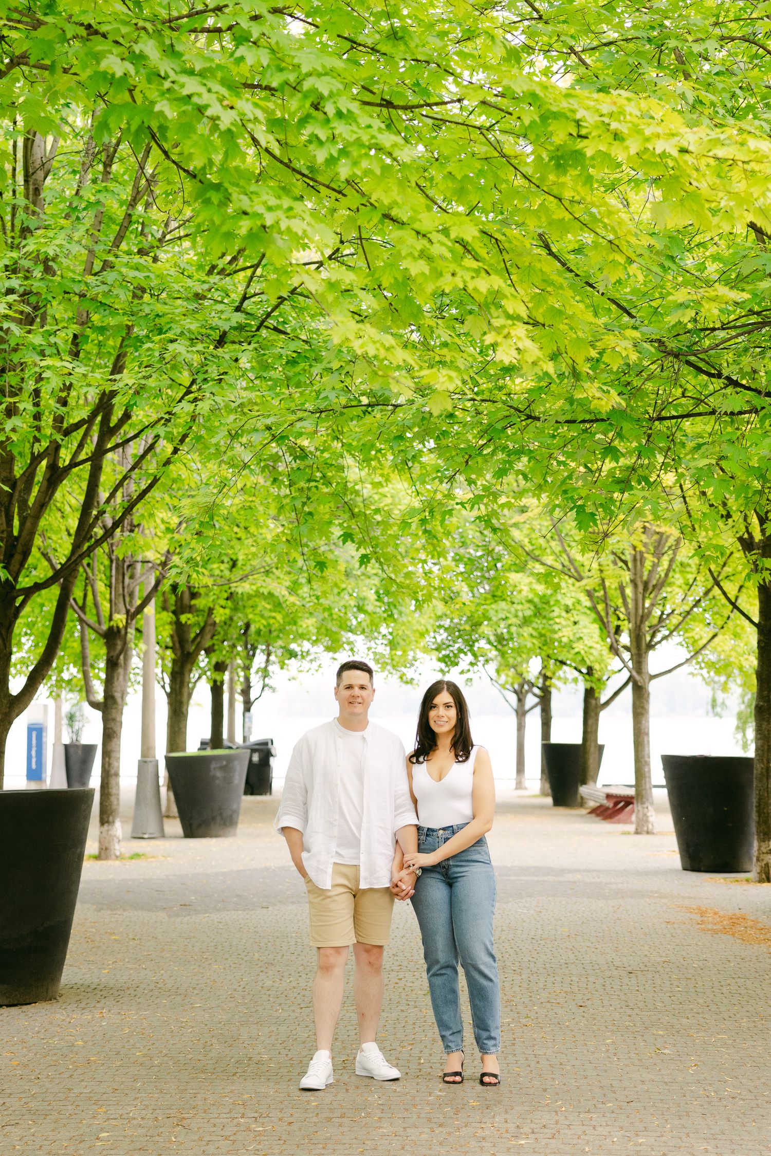 A couple poses together on a tree-lined path with large black planters.