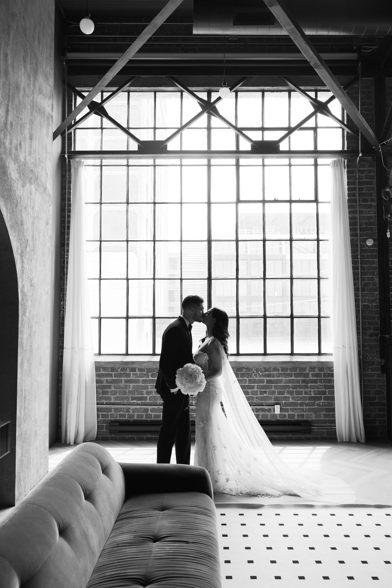 Silhouetted wedding couple stands before large industrial windows in black and white photo.