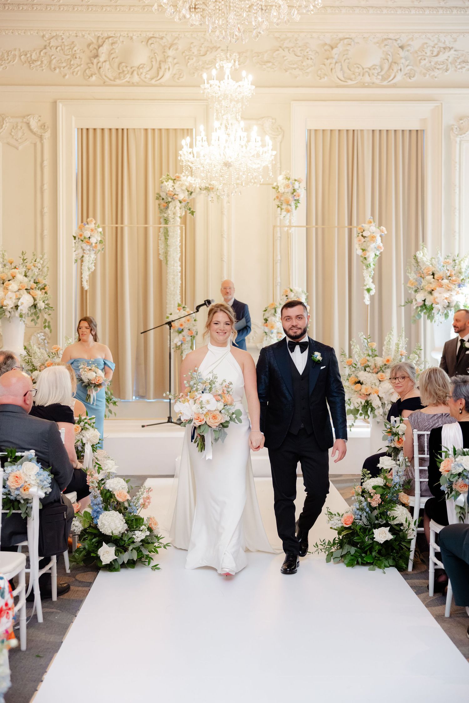 Wedding ceremony in progress in an elegant white ballroom with floral decorations.