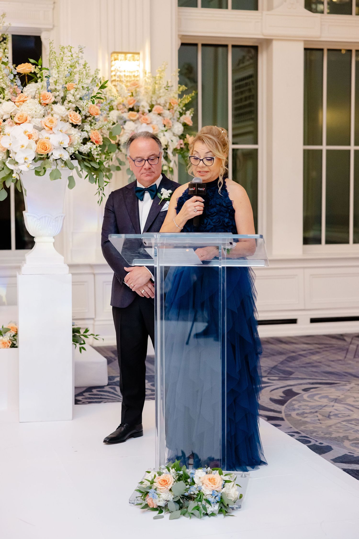 A formal event features speakers at a clear podium with elegant floral arrangements and decorative elements.
