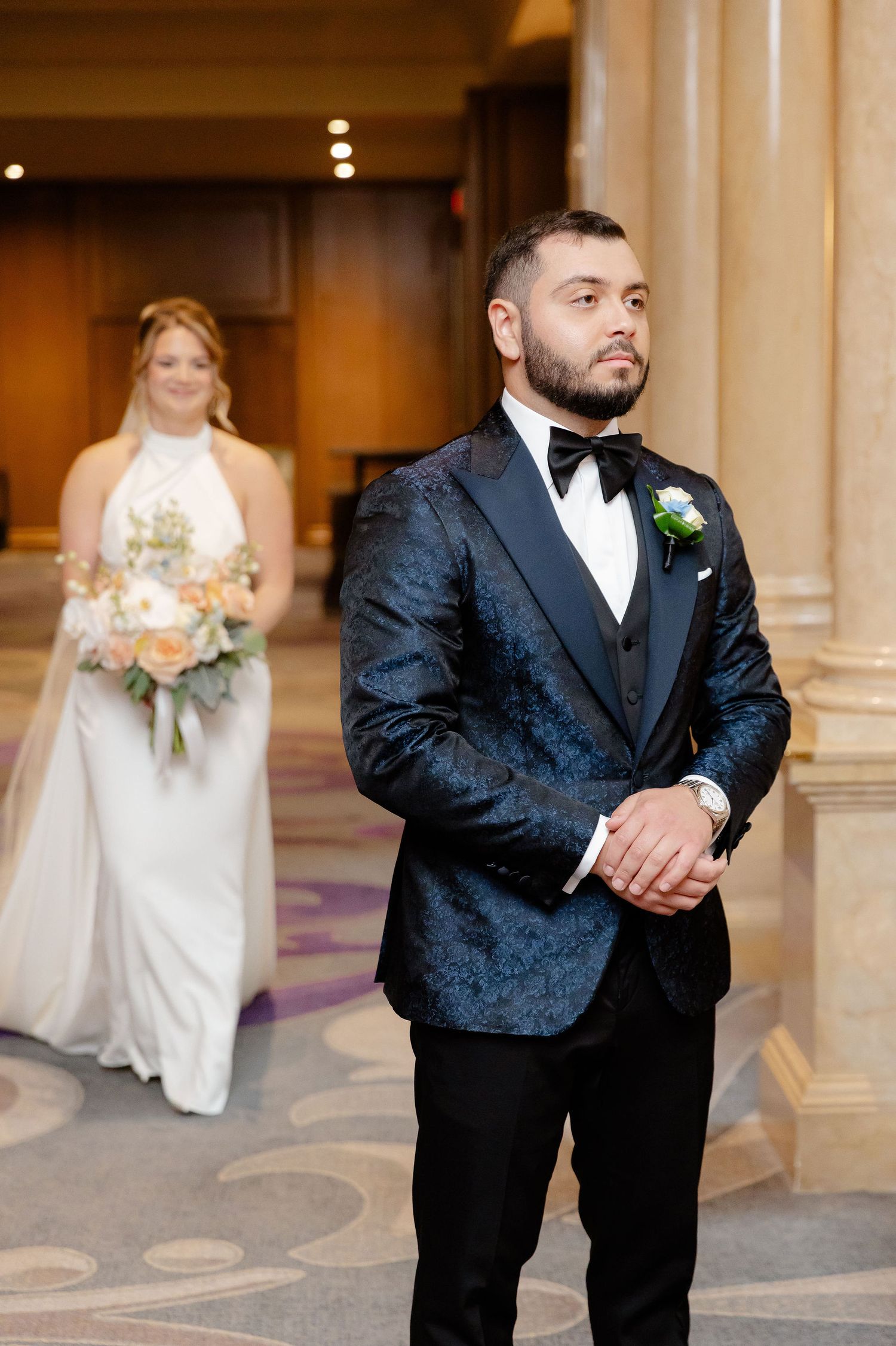 Groom in black textured tuxedo poses in an elegant venue with ornate columns.