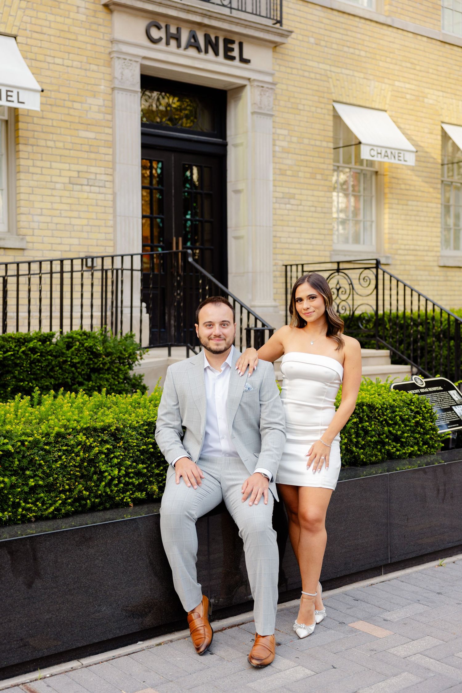 Elegant couple seated on a planter outside a Chanel boutique in coordinated formal wear.