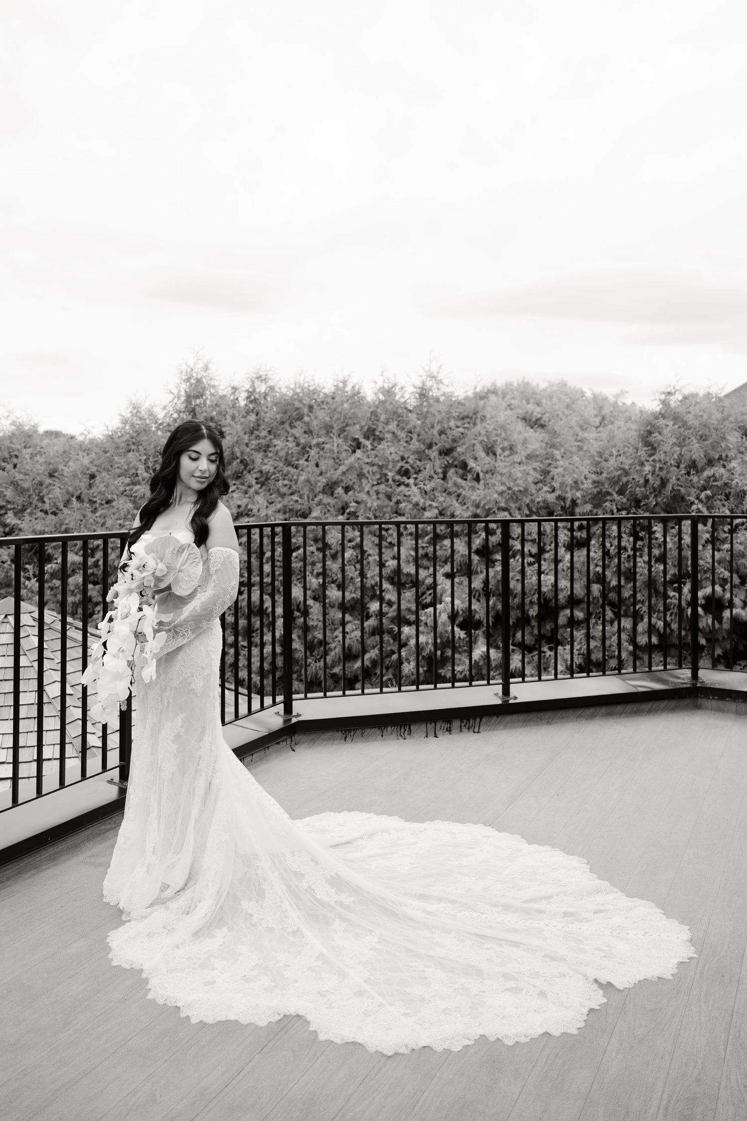 A bride in a flowing white lace gown with a long train poses elegantly on an outdoor terrace overlooking nature.