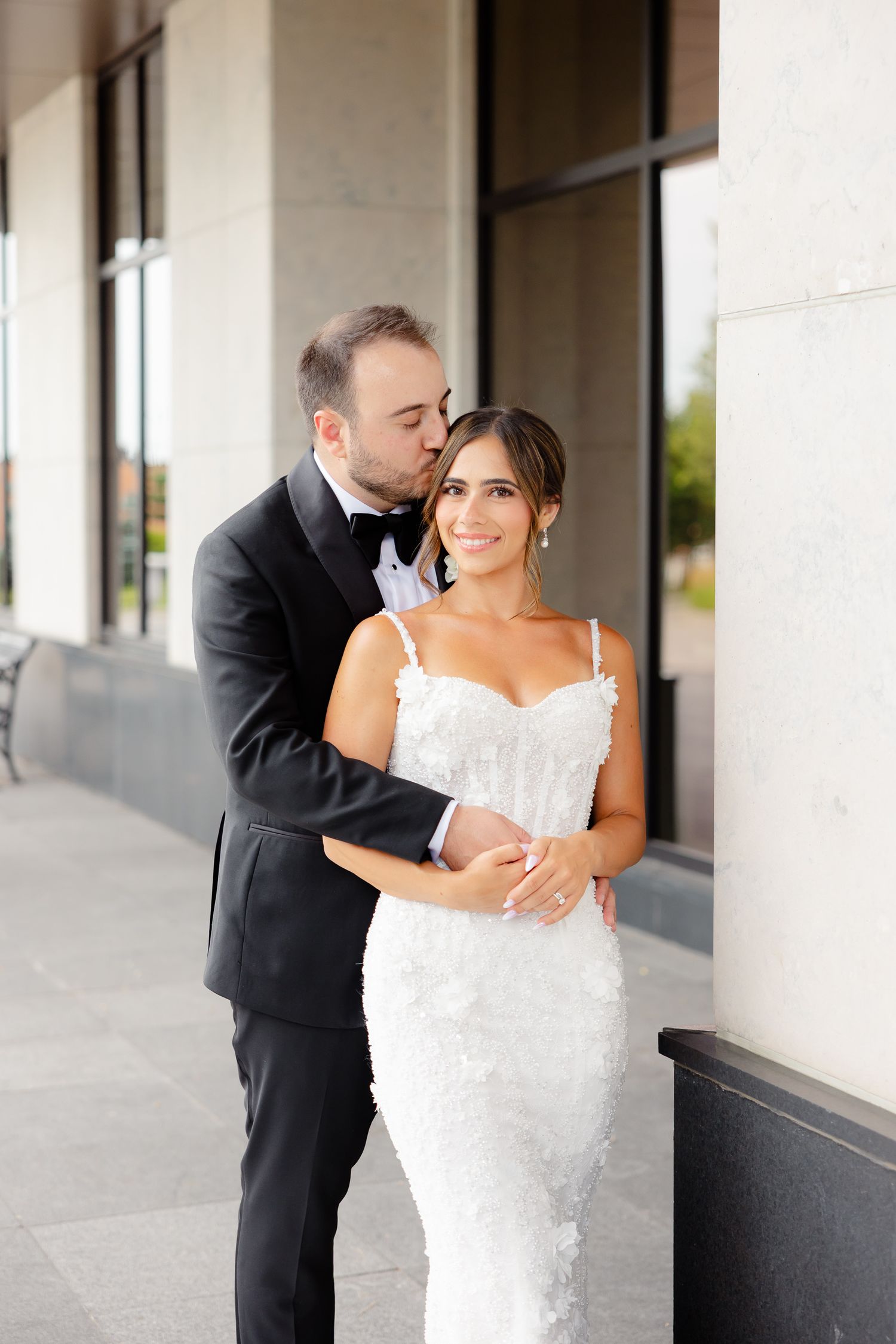 A romantic wedding couple embraces outside a building, with the bride in white lace and groom in classic black tuxedo.