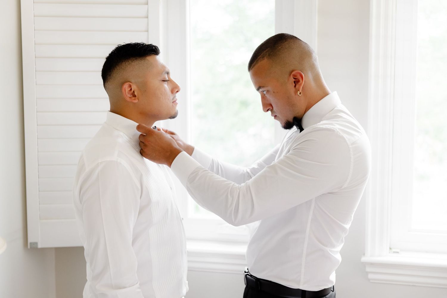 A groomsman adjusts a bowtie and boutonniere on another person at a formal event in a bright white room.