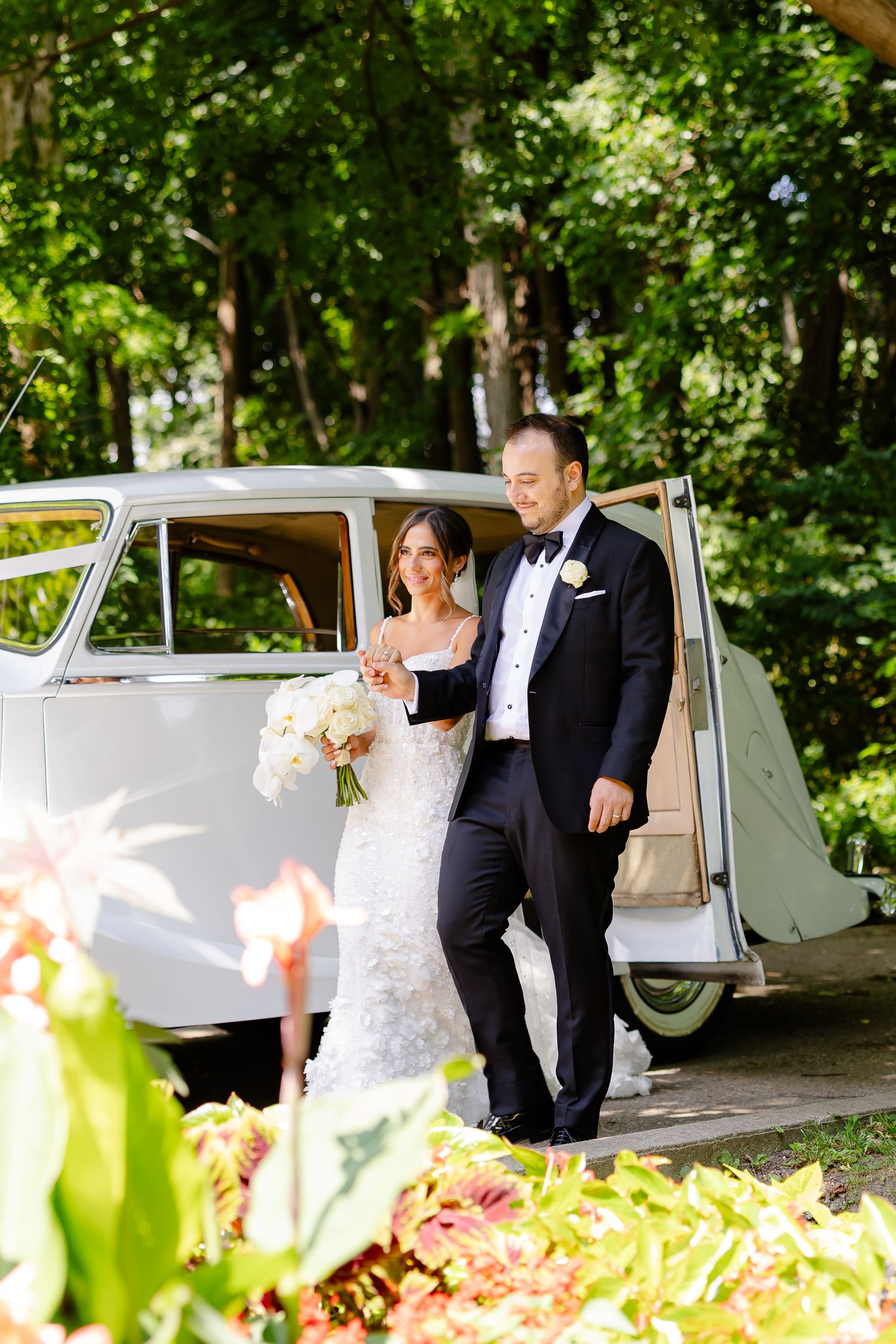 Wedding couple shares romantic moments near vintage car in beautiful garden setting with soft blooms in foreground.
