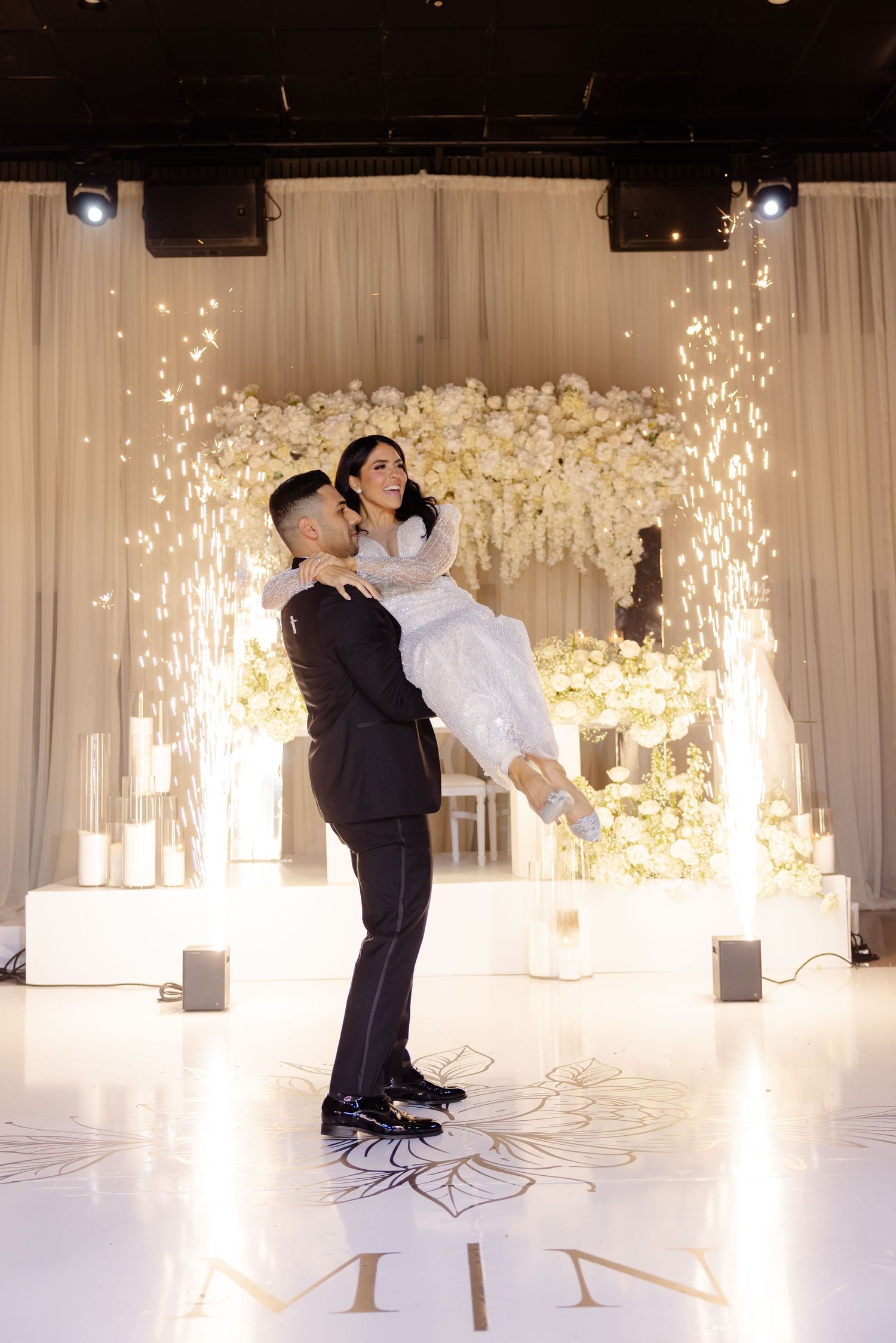 Dramatic wedding lift pose with sparklers and white floral backdrop on custom monogrammed dance floor.