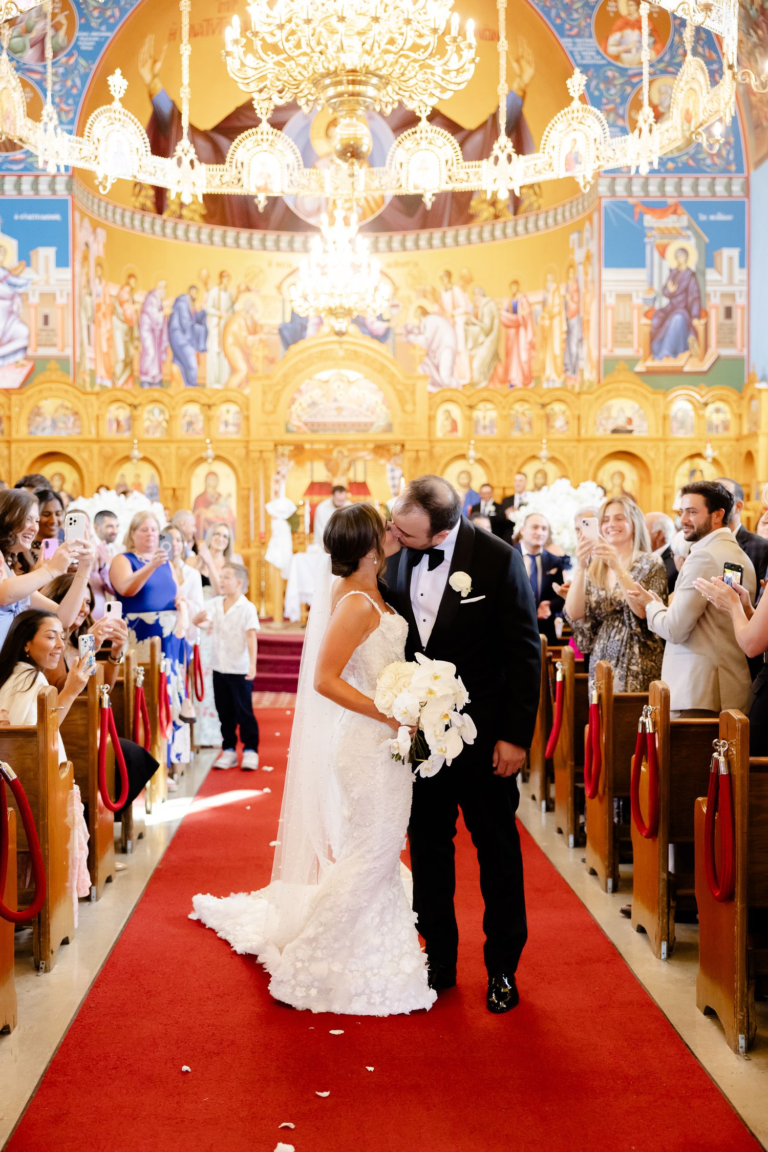 Wedding ceremony in an ornate Orthodox church with red carpet aisle and elaborate gold and blue frescoed walls and ceiling.