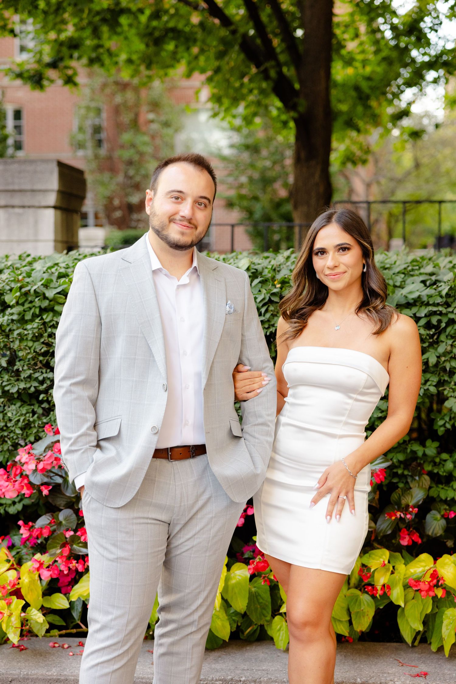 A couple in coordinated light-colored outfits poses in front of a landscaped garden setting.