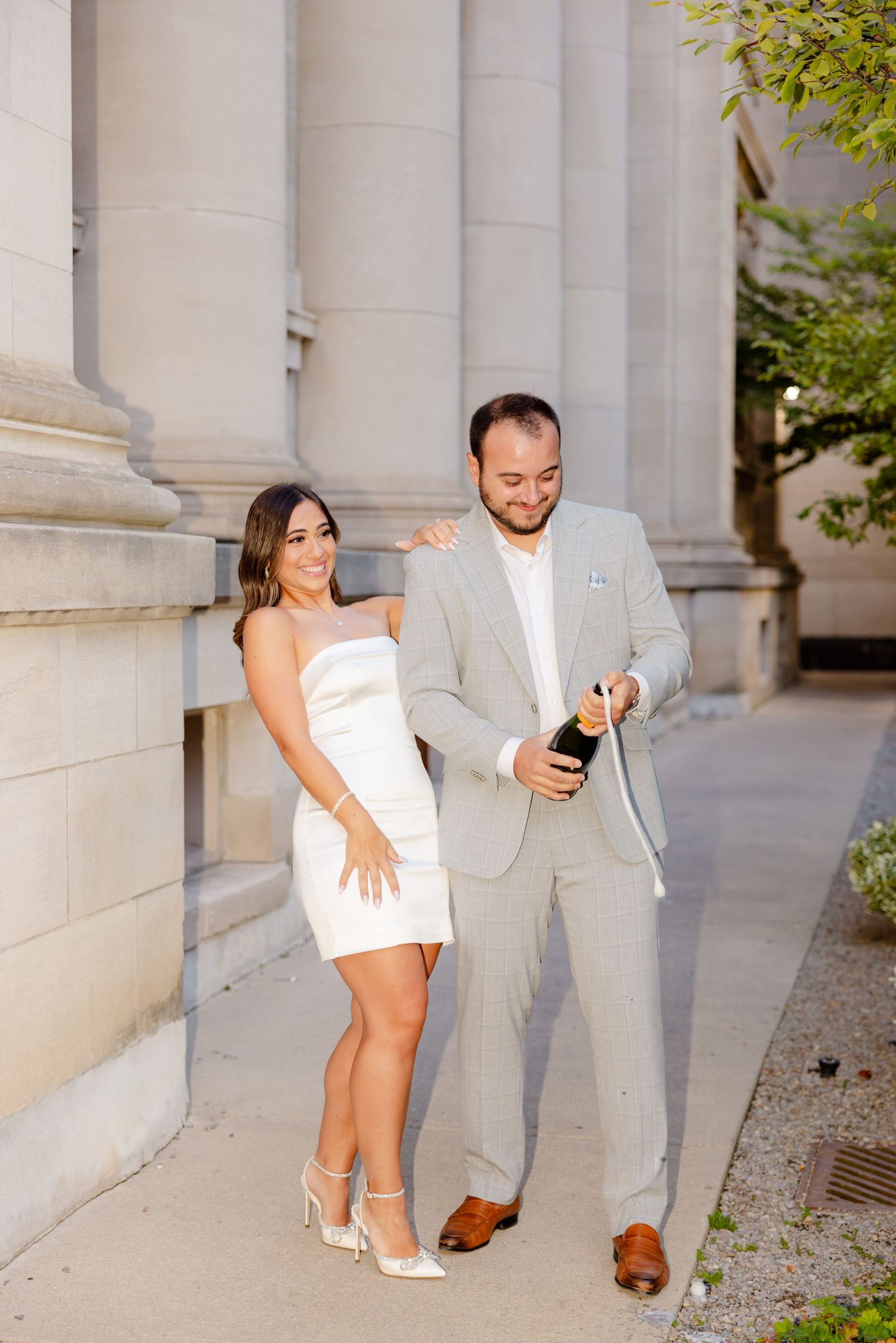 Elegantly dressed couple stands by classical columns while holding champagne bottle.