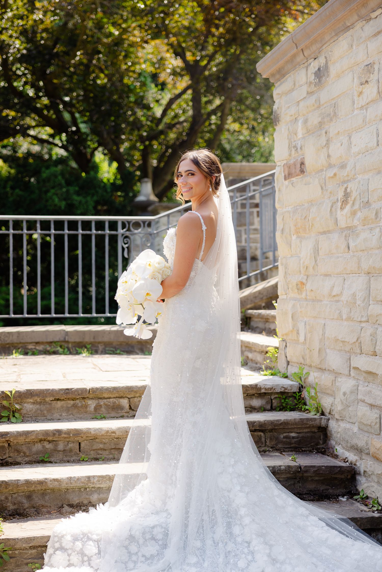 A bride in a flowing lace wedding gown with train stands on stone steps holding a white bouquet on a sunny day.