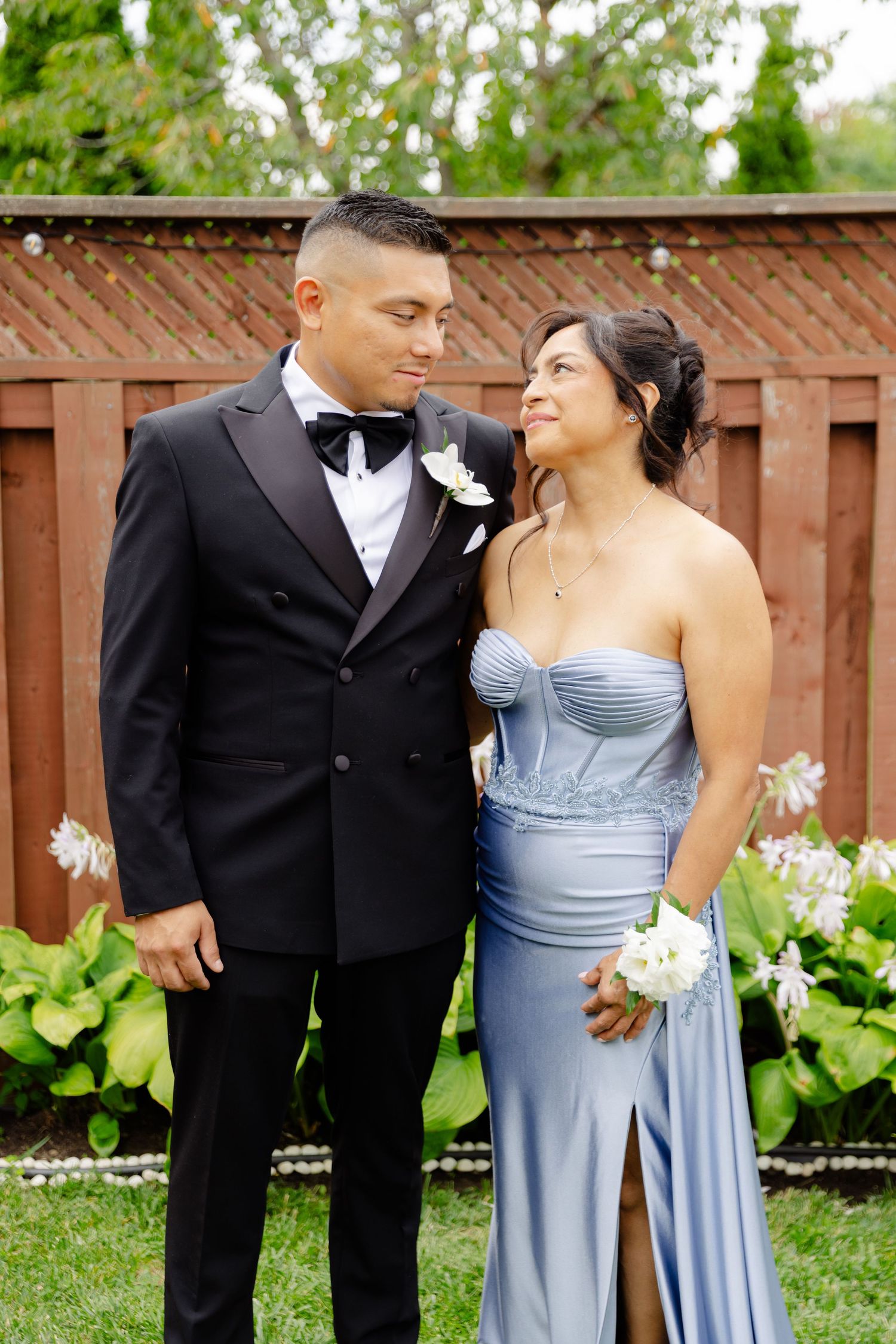 A couple in formal wedding attire stands in front of a wooden fence, the bridesmaids wearing light blue dresses.