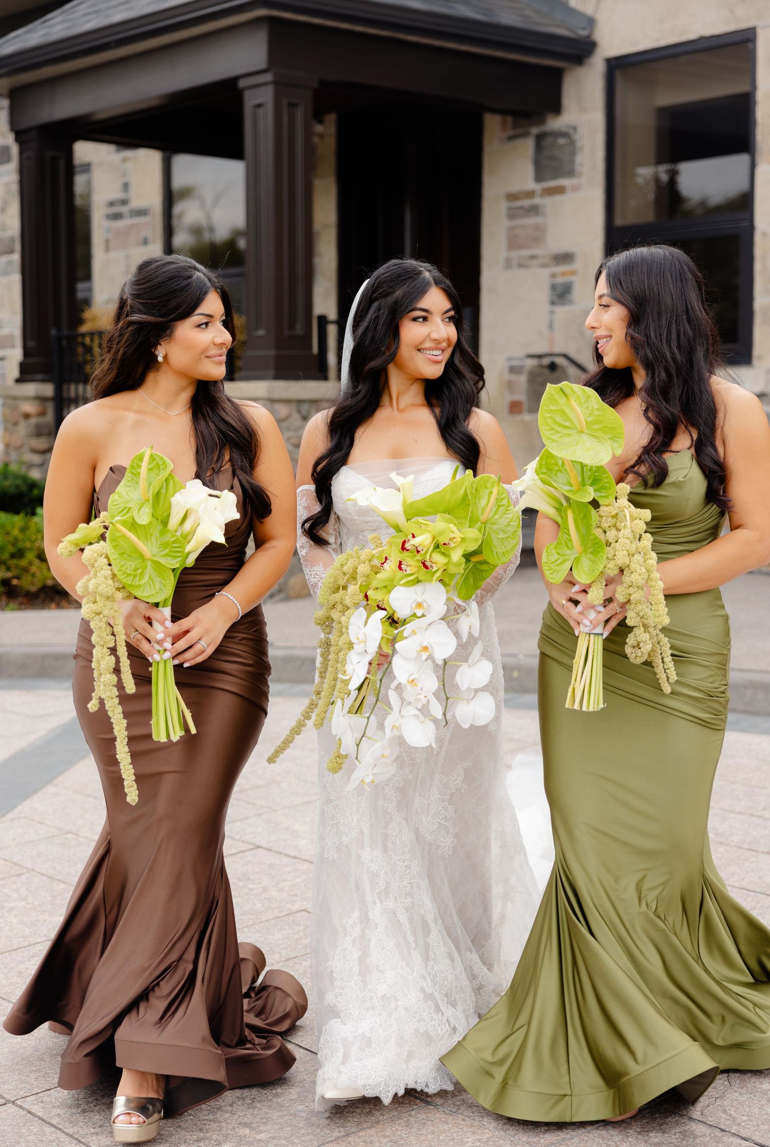 Three women in elegant gowns hold unique green and white floral bouquets outside a building entrance.