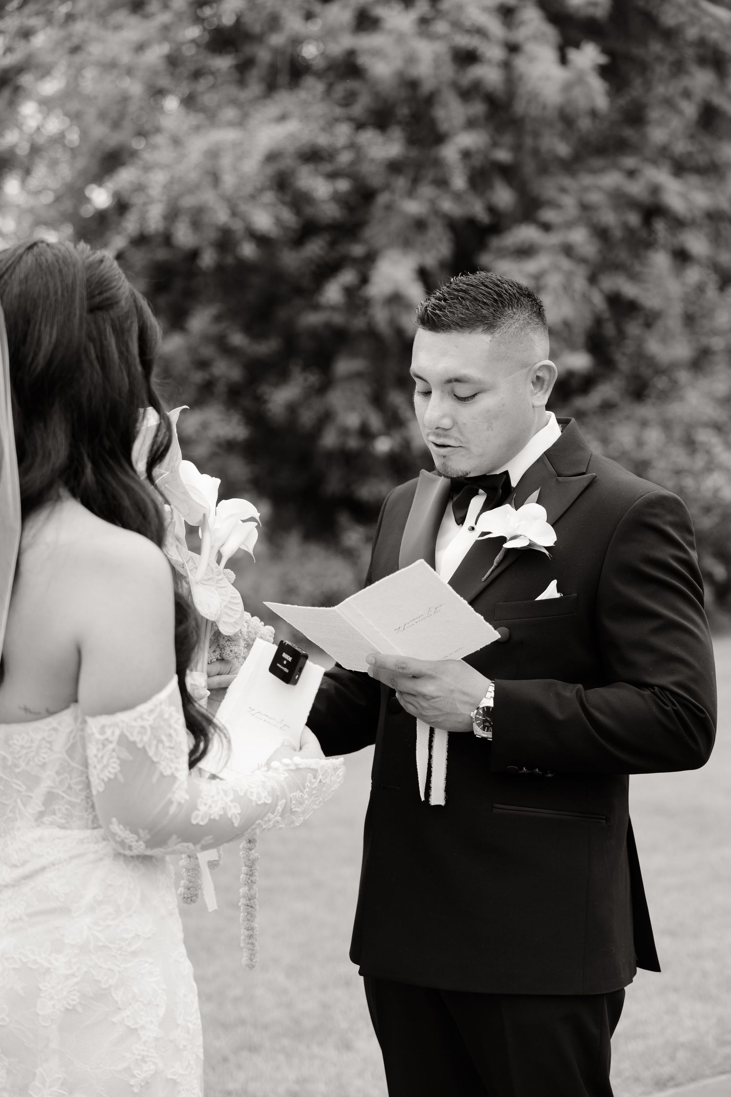 A black and white wedding photo showing a groom in a tuxedo reading vows with an emotional bride during their ceremony.