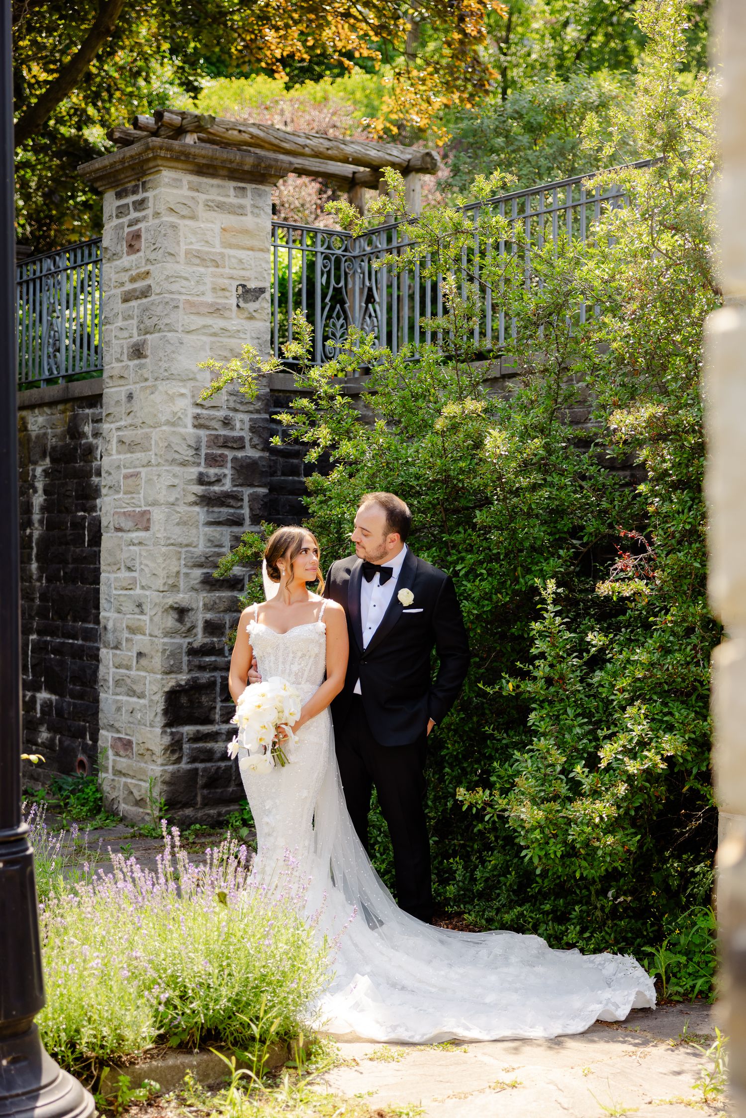 A couple in wedding attire stands by a stone wall and wrought iron fence surrounded by lush garden greenery.