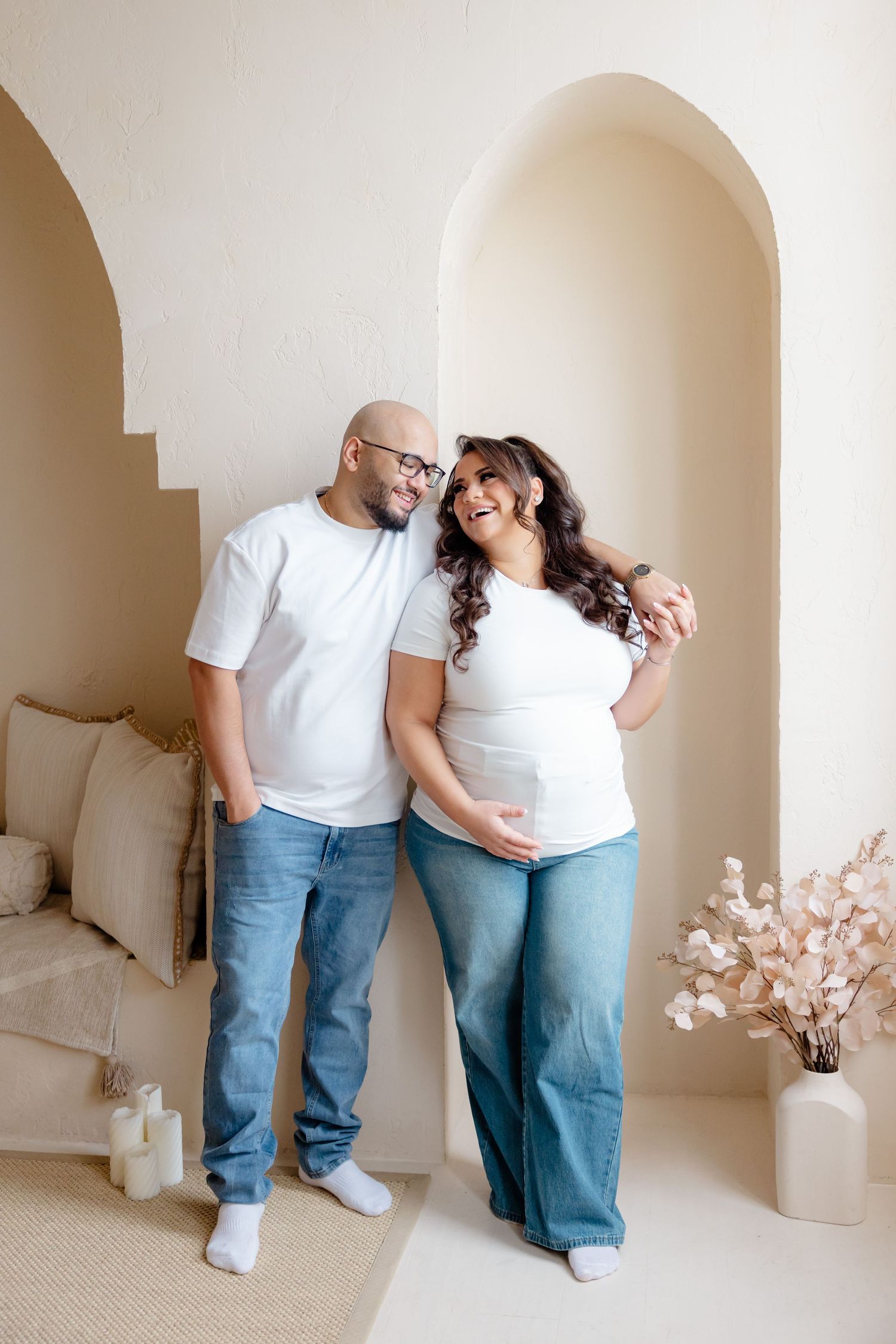A couple in matching white t-shirts and jeans embrace while standing in an arched doorway with minimalist decor.