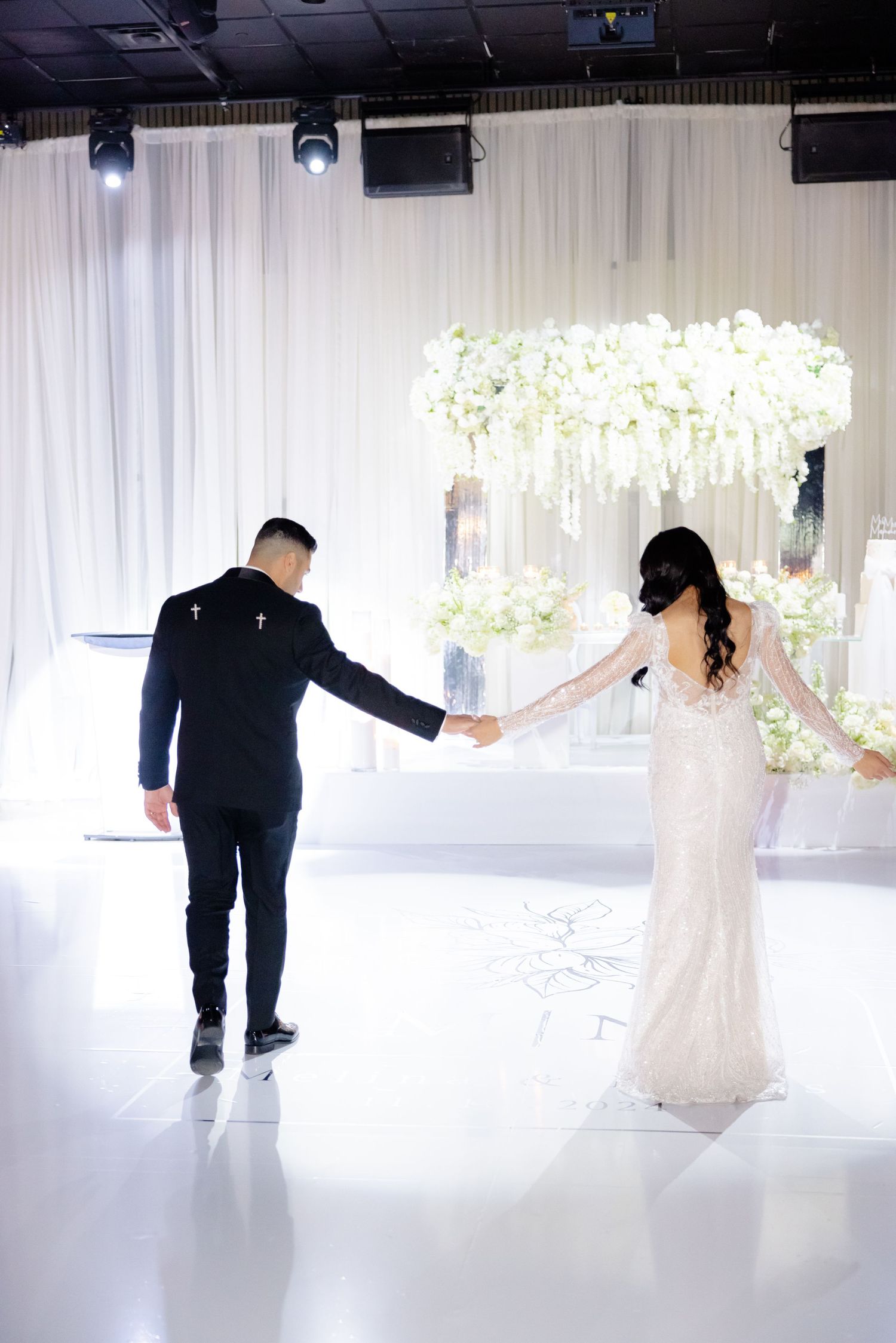 A couple walking hand in hand beneath a luxurious white floral arch in an elegant white venue.