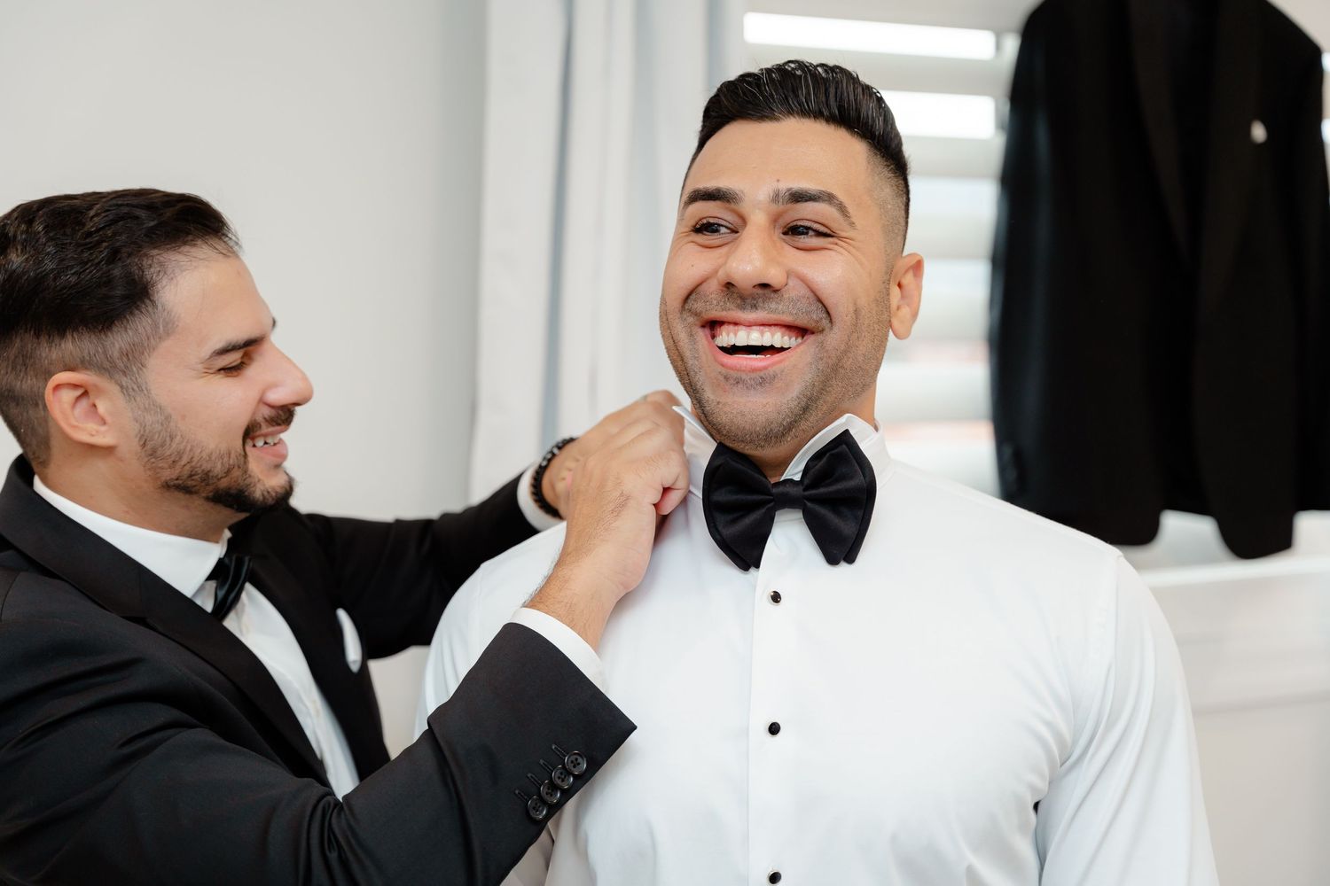 Best man adjusting groom's bow tie while sharing joyful laughter during wedding preparations.