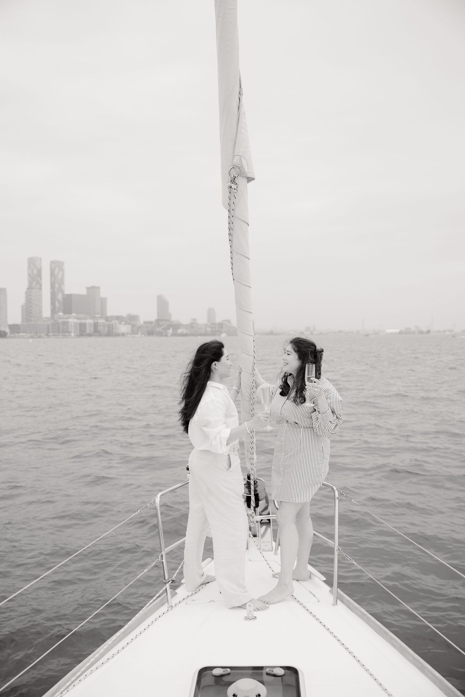 Two women stand on the bow of a sailboat, looking out at a city skyline across the water on a hazy day.