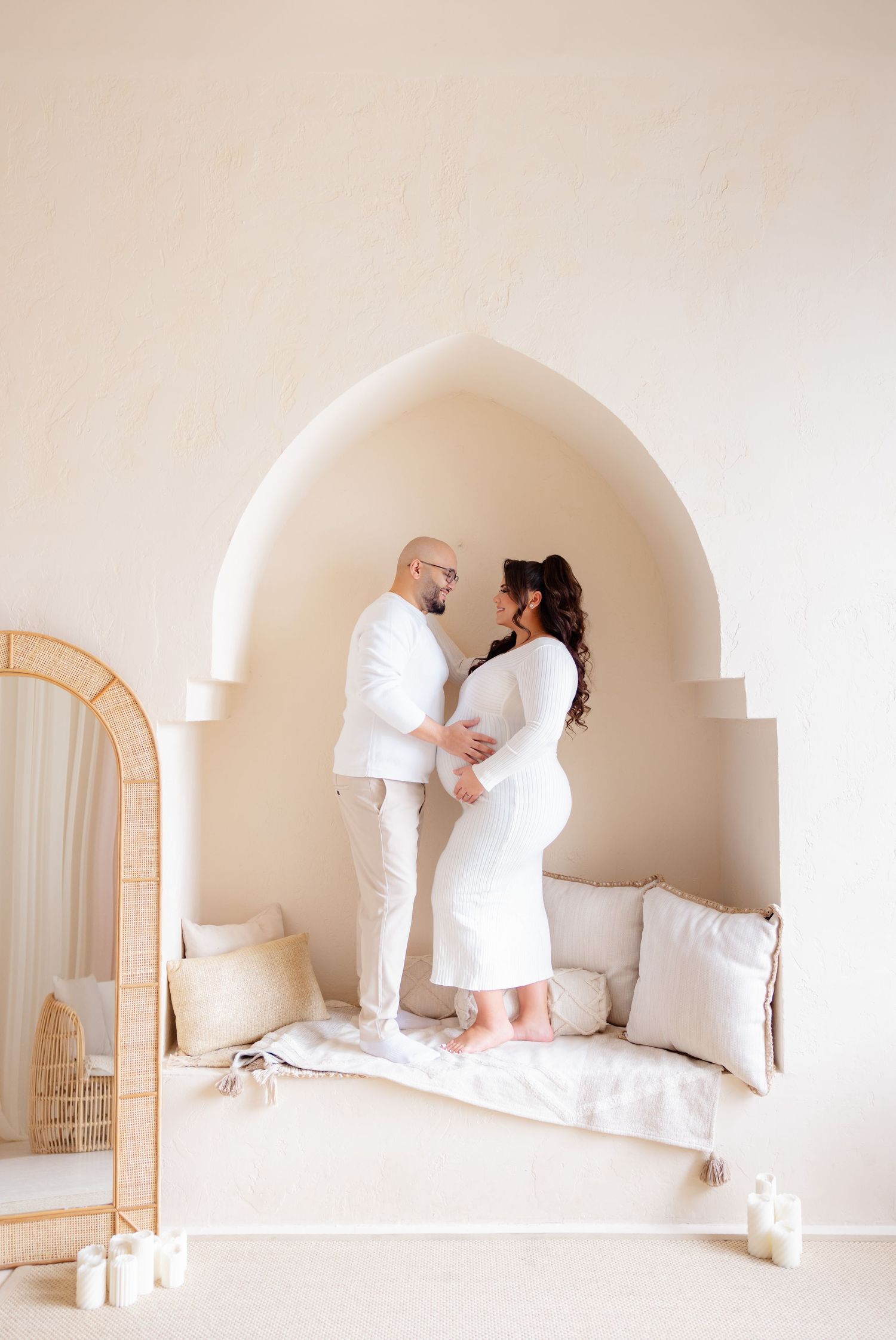 Couple in all-white outfits standing in an arched alcove with neutral decor and candles on the floor.
