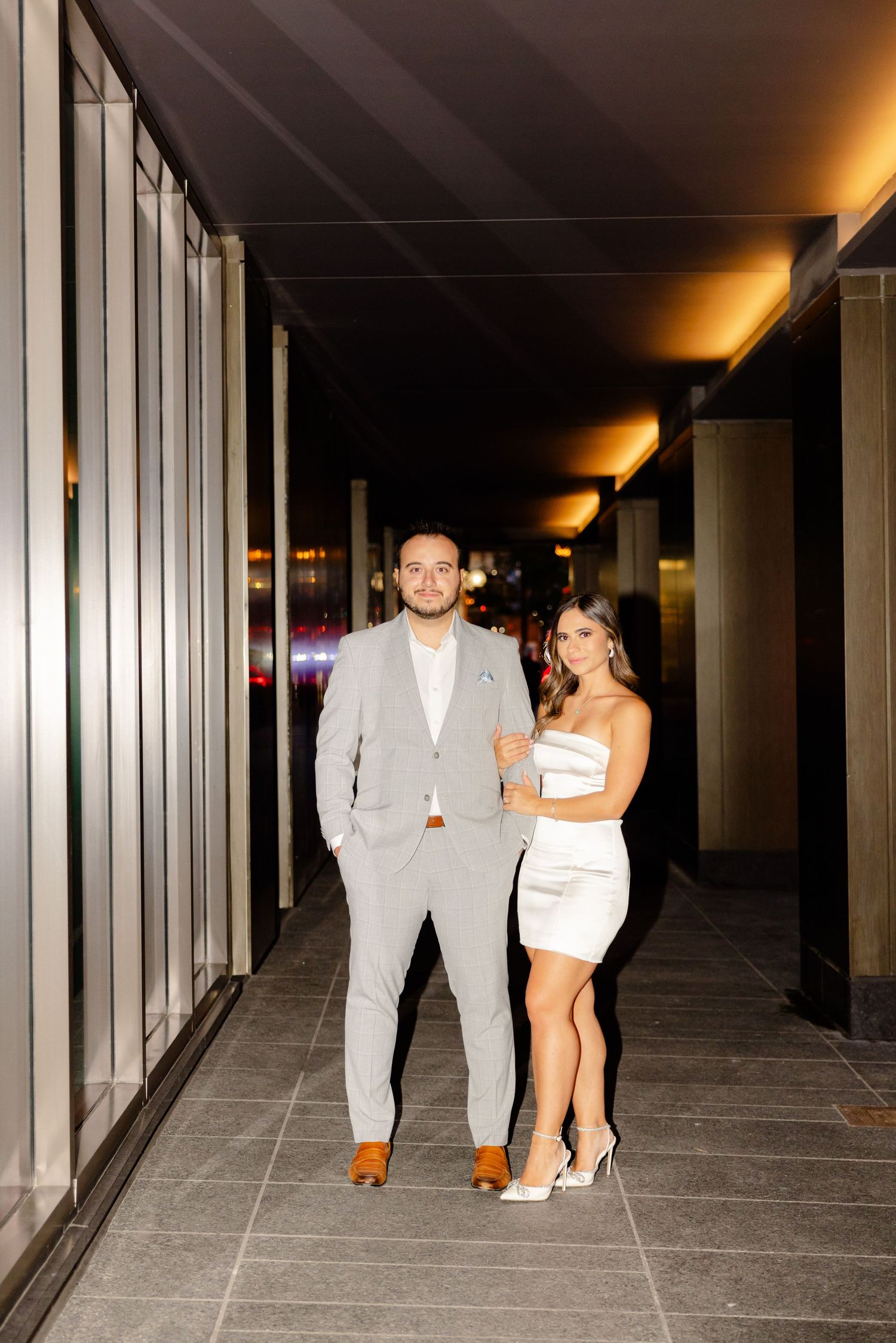 Well-dressed couple posing in a modern hallway with warm ambient lighting.