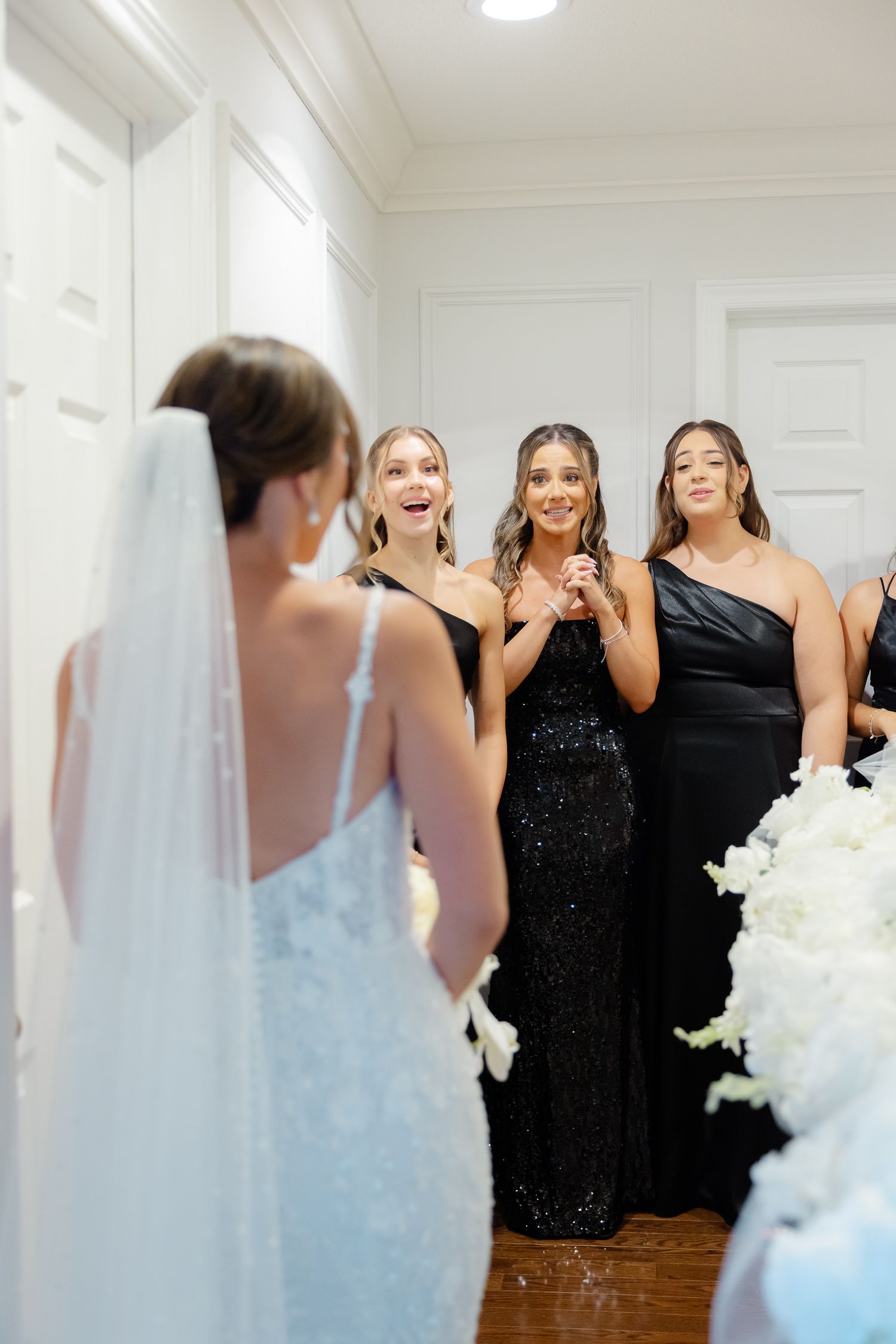 Bridesmaids in elegant black dresses react with excitement and joy upon seeing bride in her wedding gown and veil.