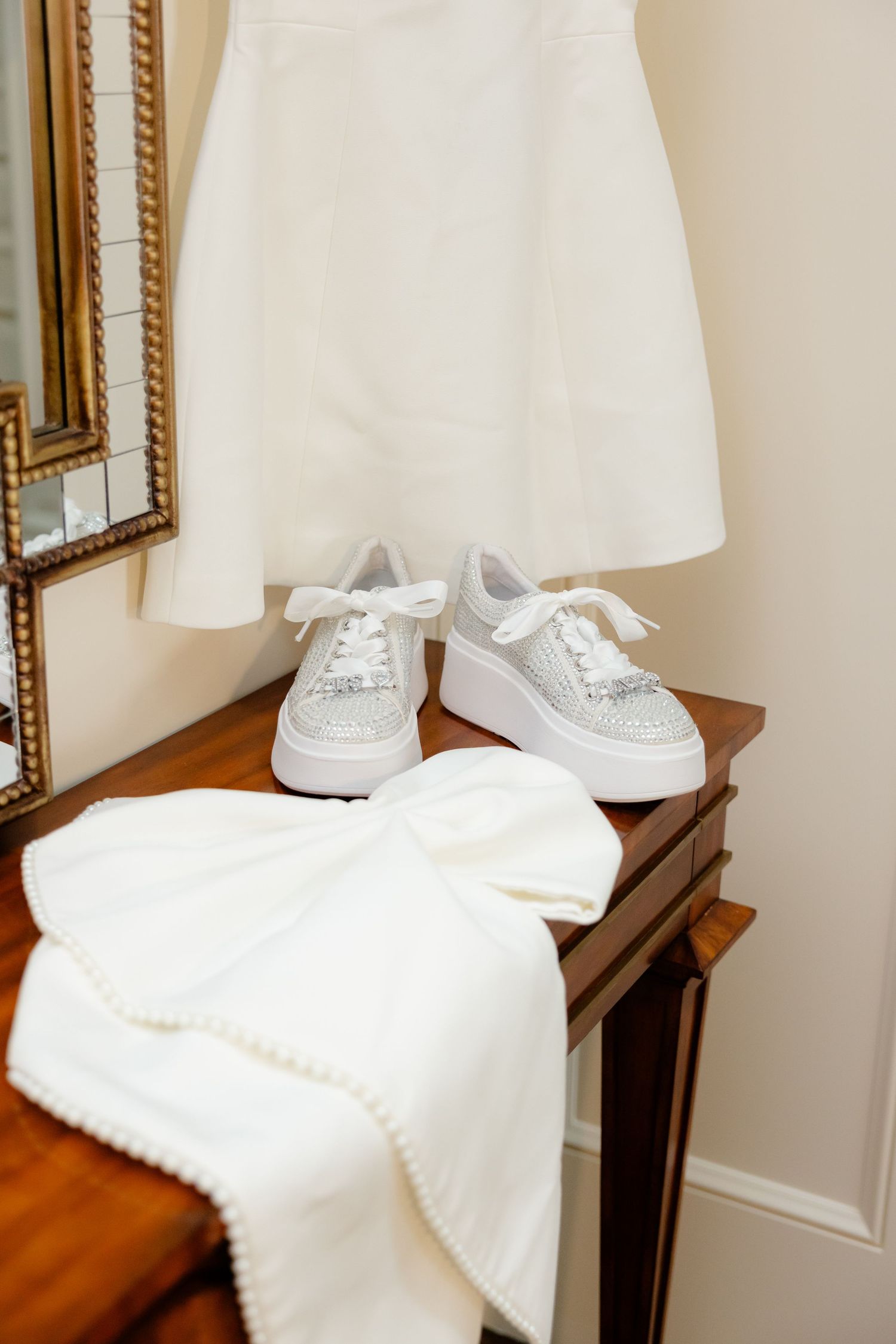 White sparkly sneakers and wedding dress displayed on wooden console table beside ornate gold mirror.