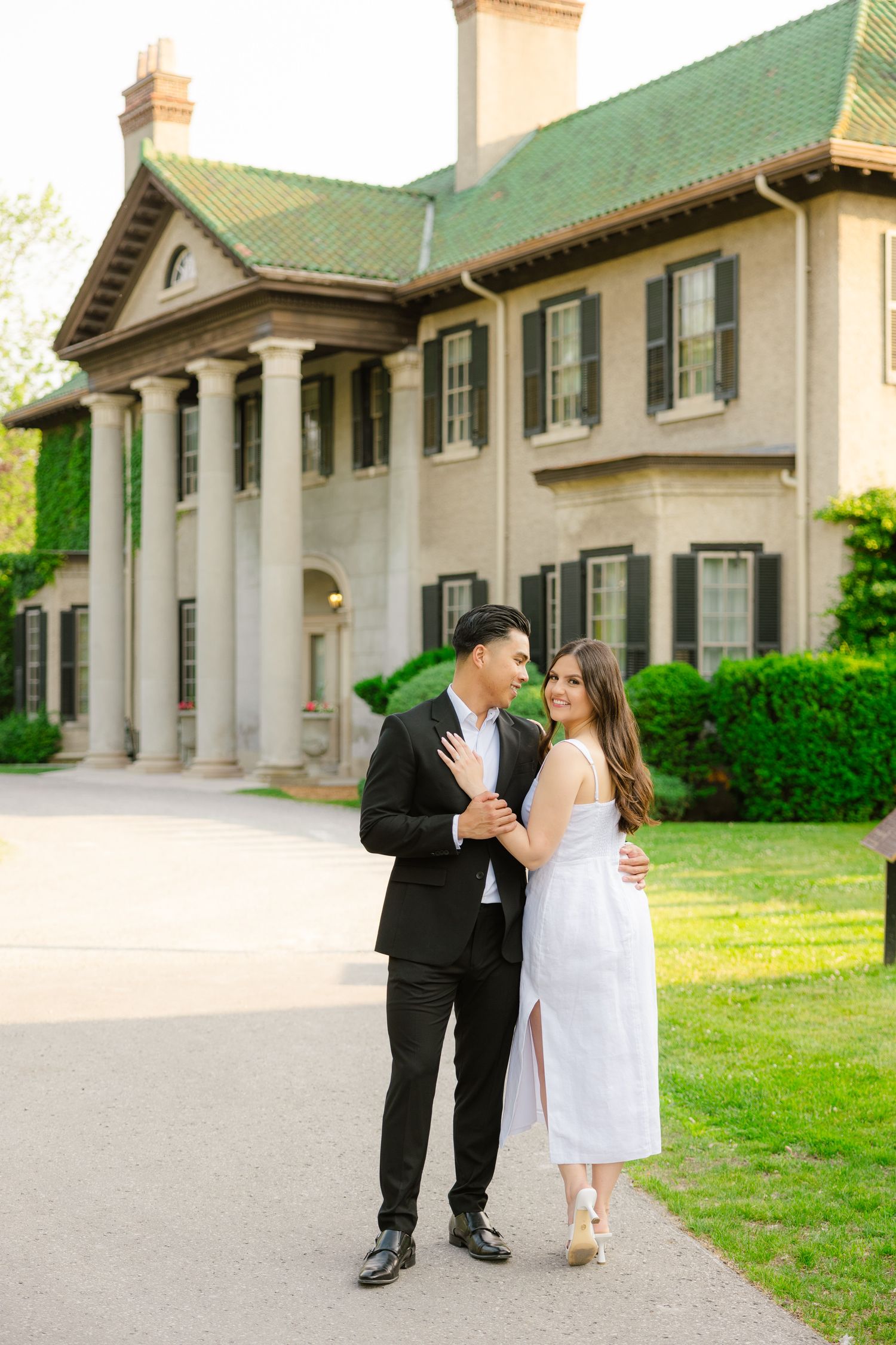 Elegant couple poses in formal attire in front of a historic mansion with white columns and green roof.