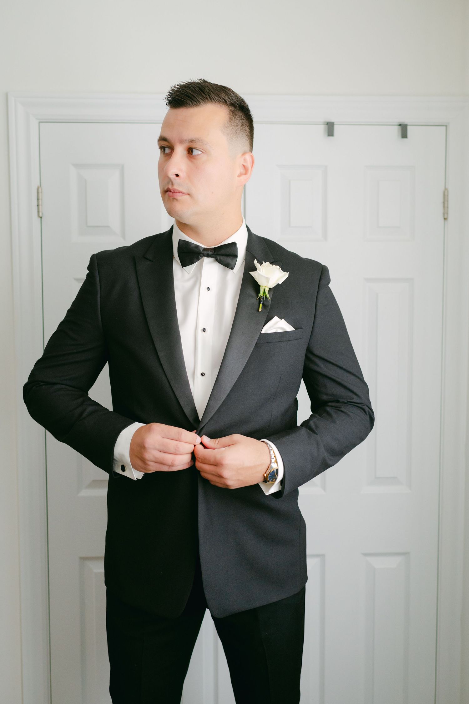 A groom in a classic black tuxedo with white boutonniere adjusts his jacket before the ceremony.
