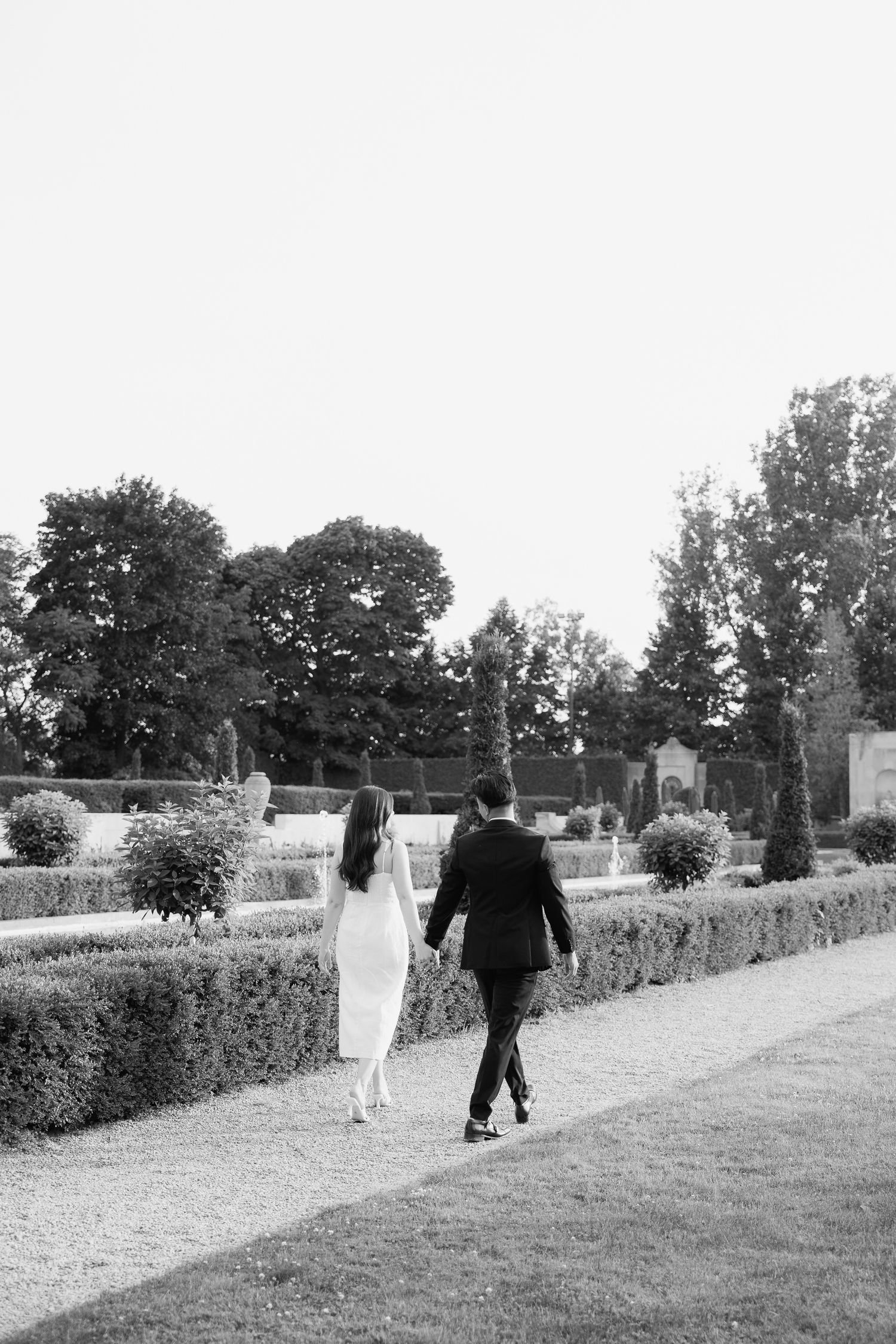 A couple walking away hand in hand through a formal garden with manicured hedges.