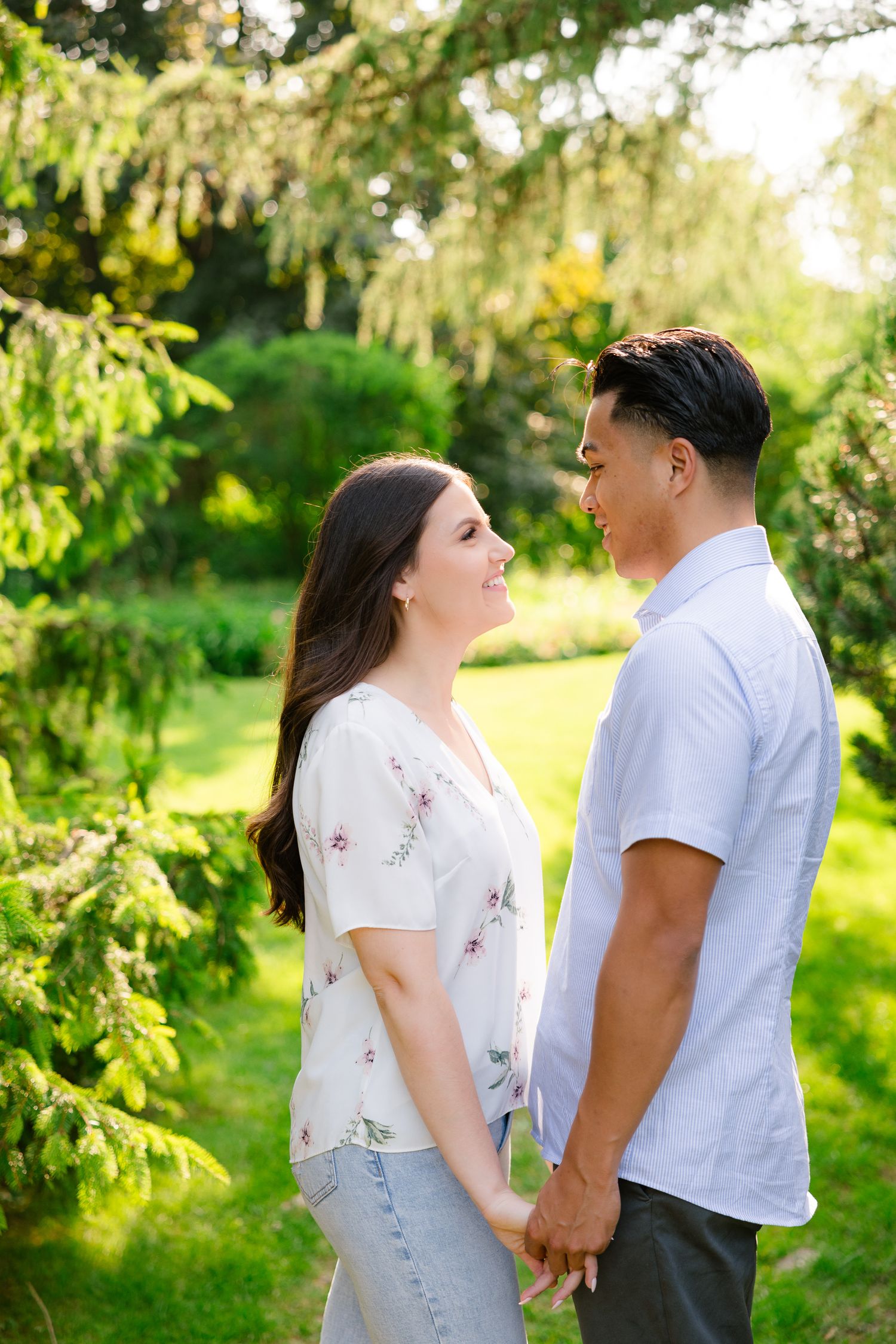 A couple shares a romantic moment while holding hands in a sunlit garden with drooping pine branches.