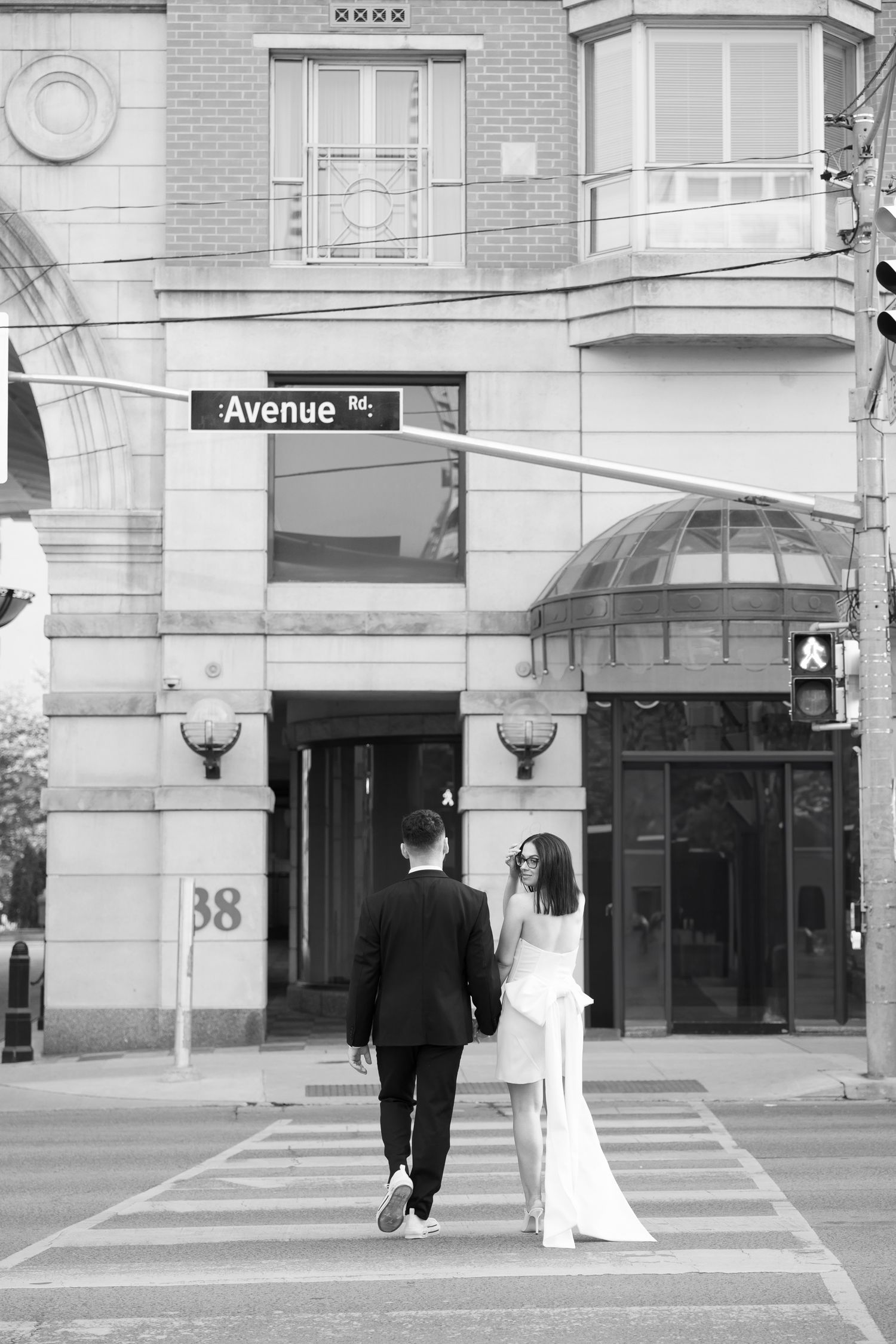A couple in formal attire walking across a crosswalk in front of a building with an Avenue street sign.