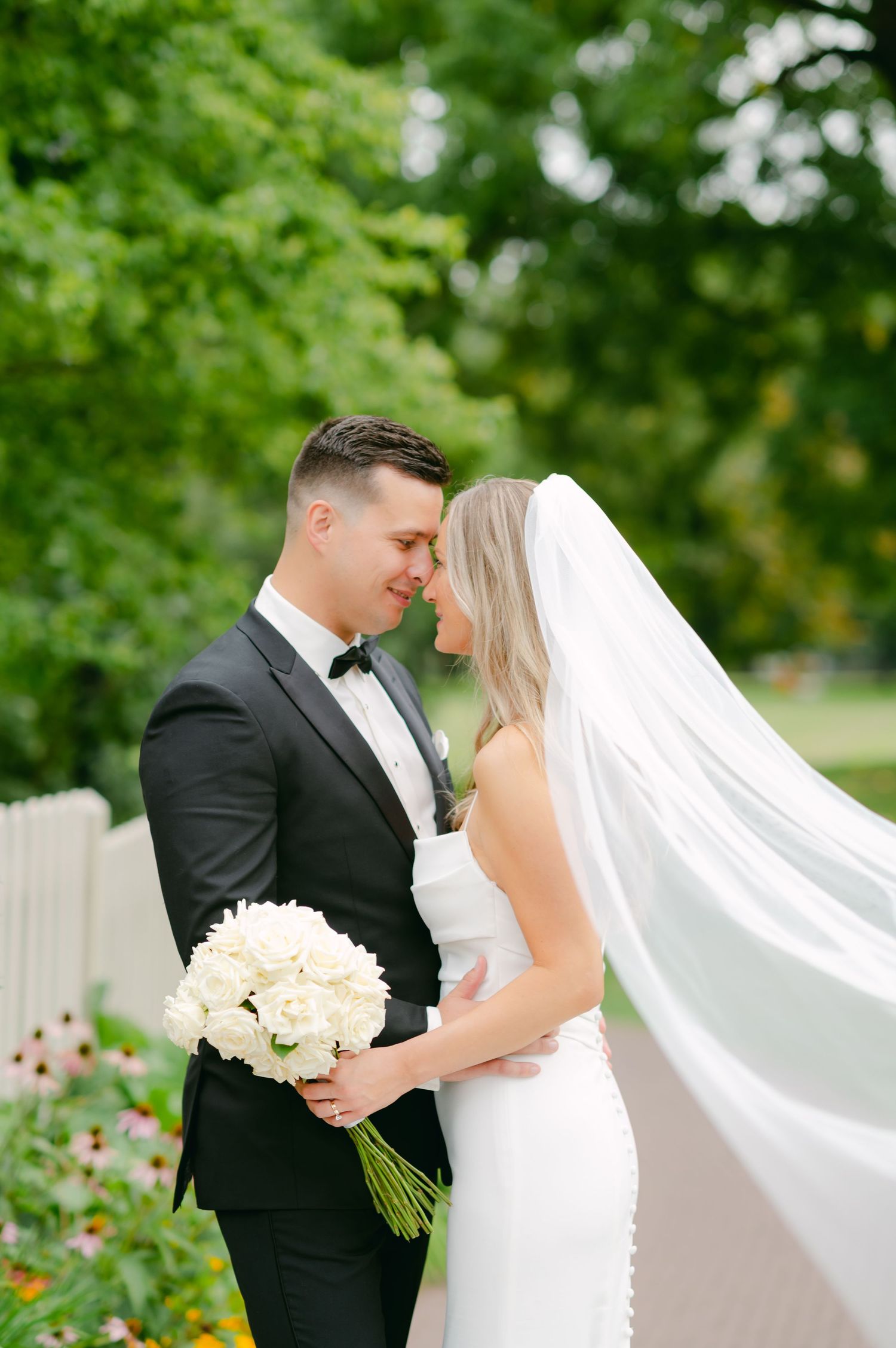 Elegant wedding couple posing outdoors with flowing veil and white roses.