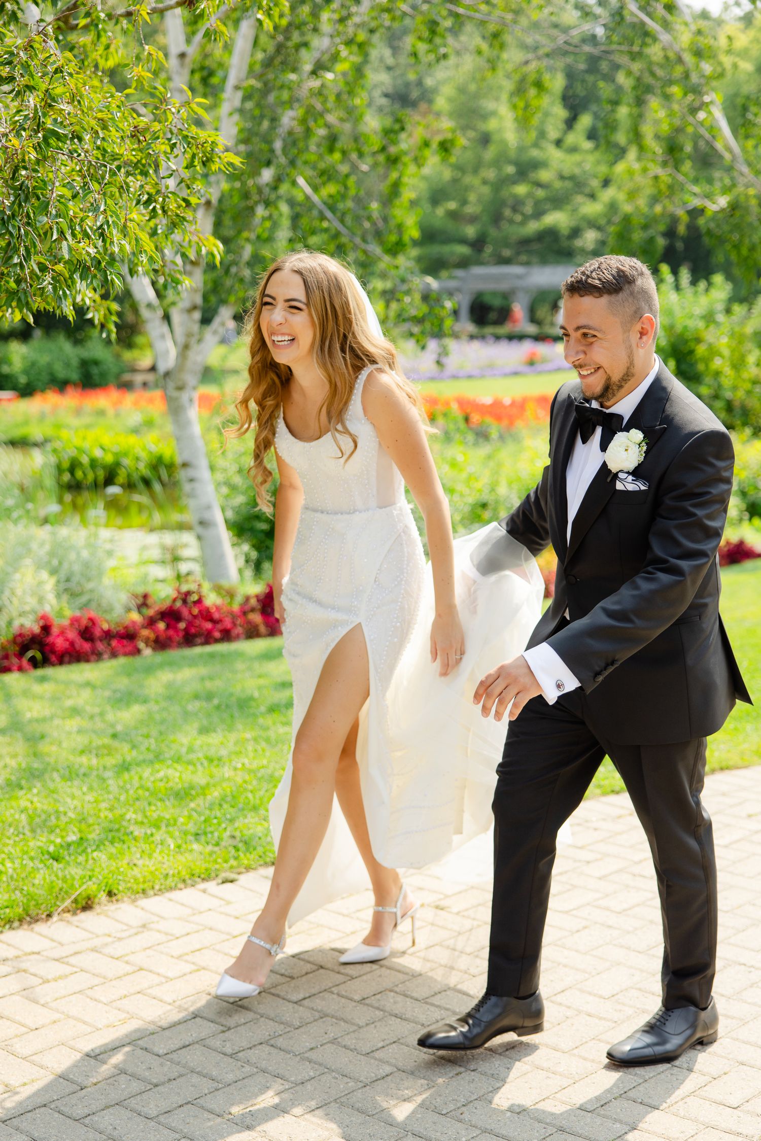 Newlyweds laughing while walking together on a garden path, with the bride in an elegant slit dress.
