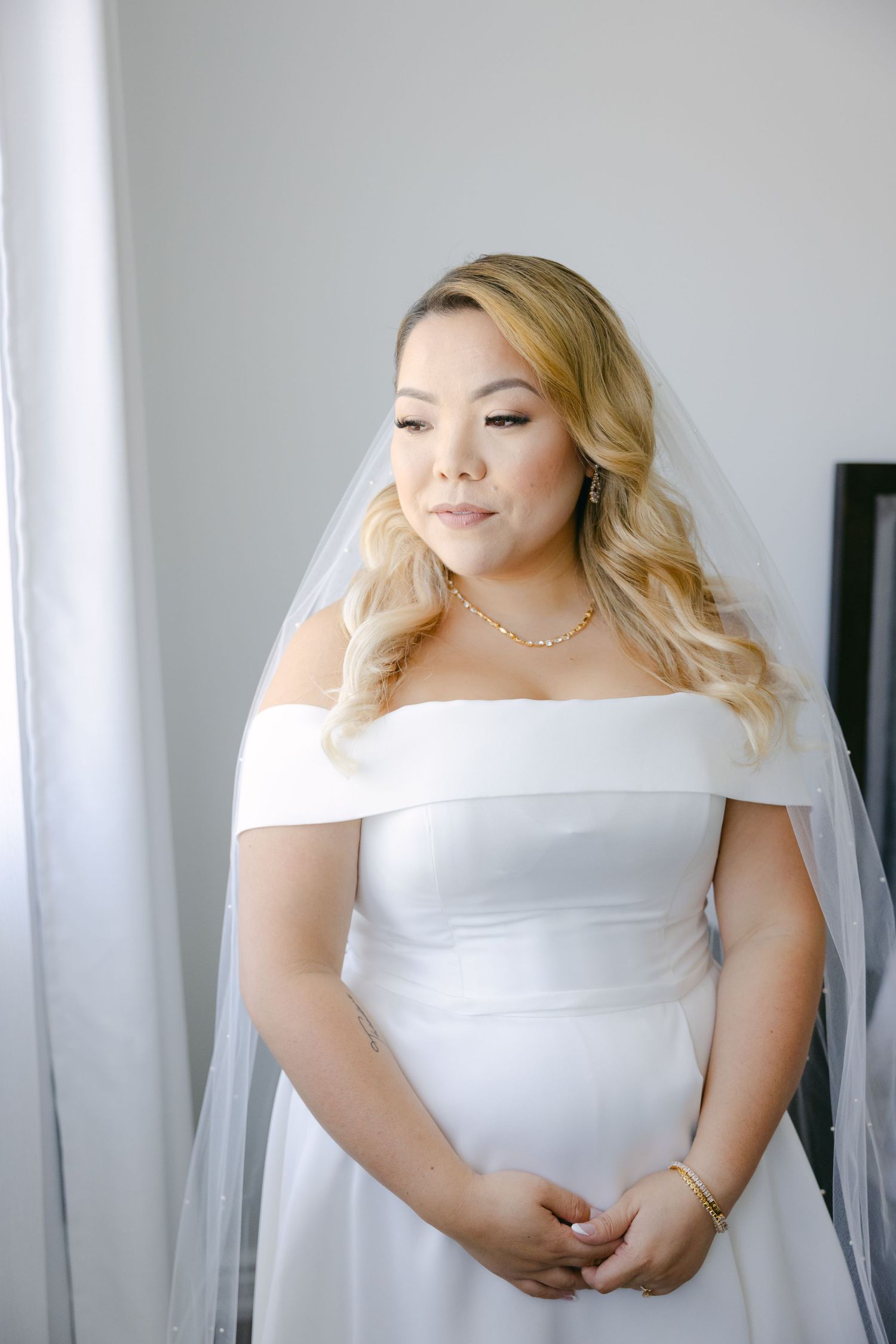 Bride in elegant off-shoulder dress and veil poses against light background.