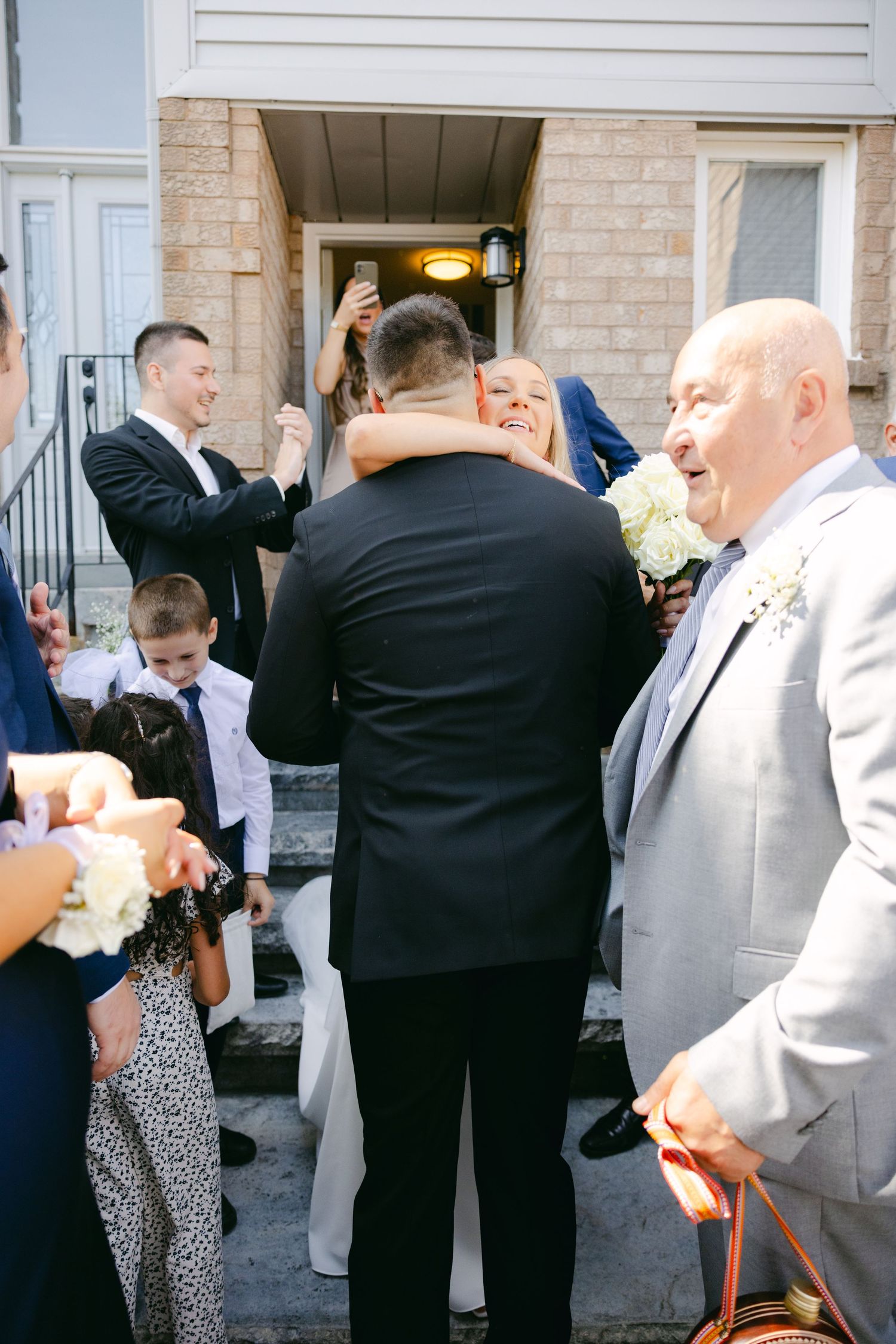 Wedding party and guests sharing joyful moments outside brick building entrance.