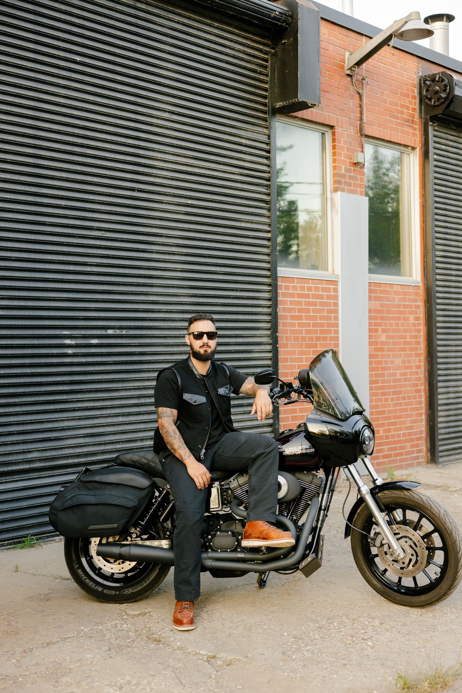A motorcyclist in casual wear poses with their black cruiser motorcycle against a striped wall exterior.