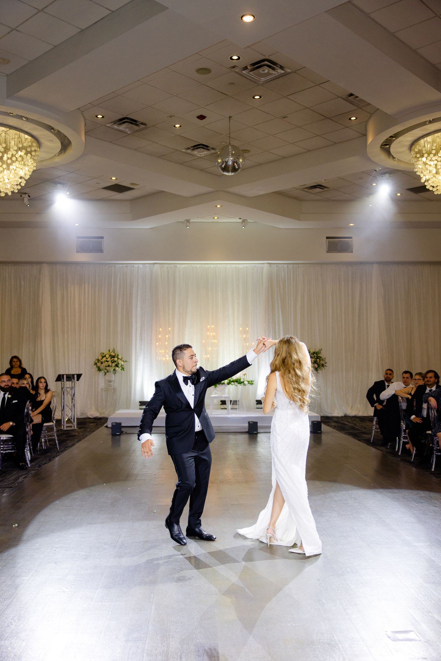 Elegant couple performs their first dance in a beautifully lit ballroom with white drapes and crystal chandeliers.