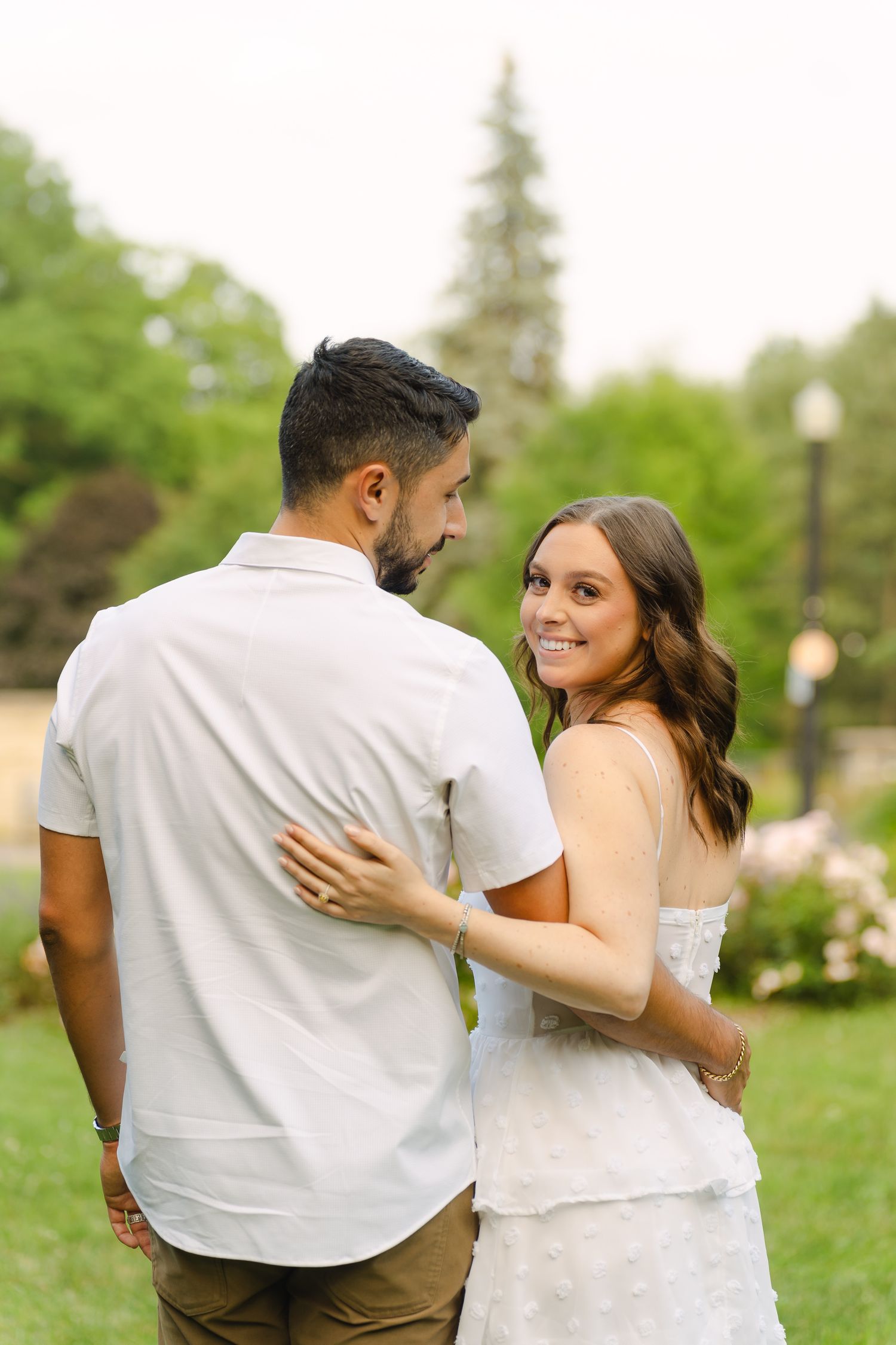 A couple shares an intimate moment in a garden with flowering bushes and trees in the background.