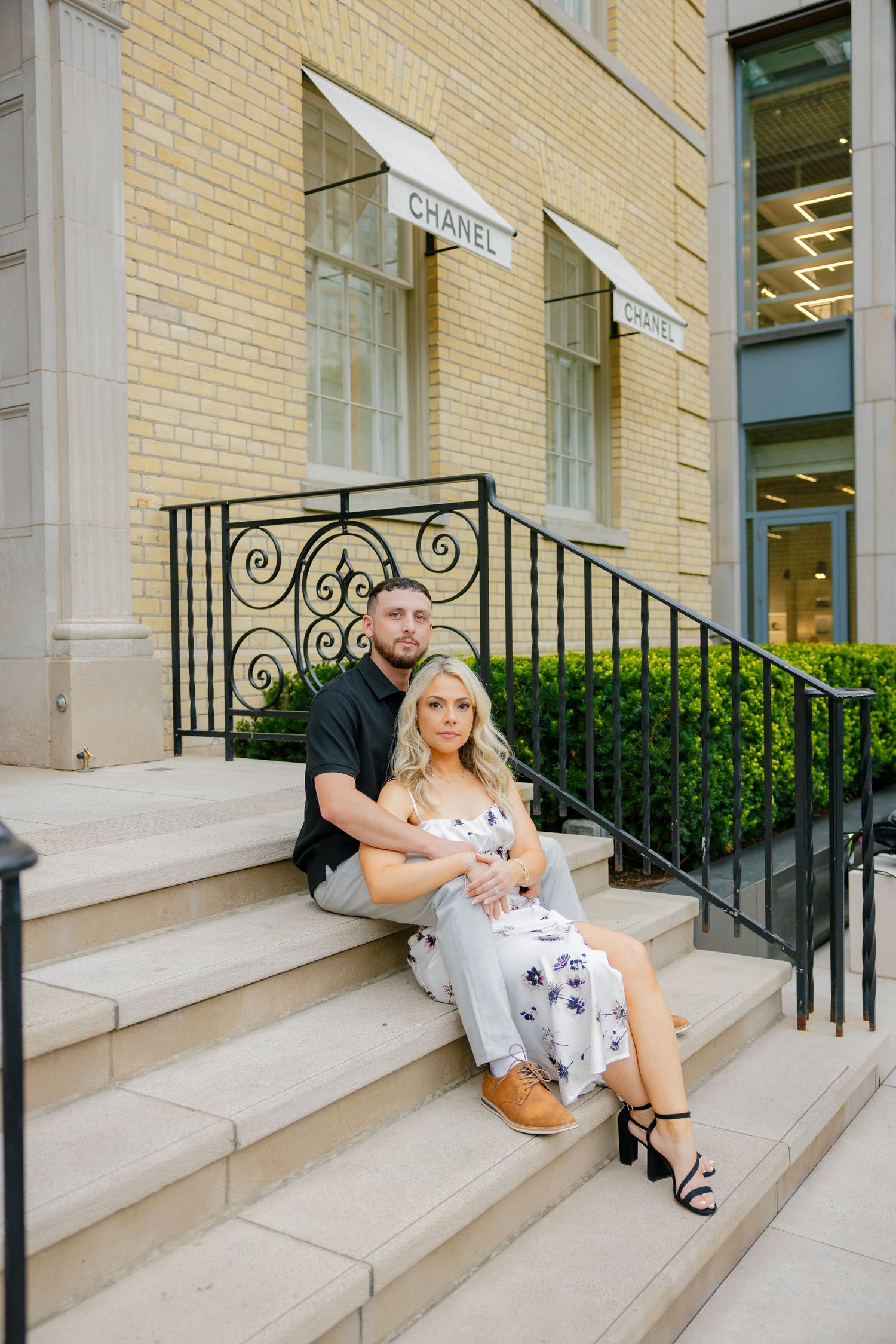A couple poses together on the steps of a luxury boutique entrance with ornate black railings.