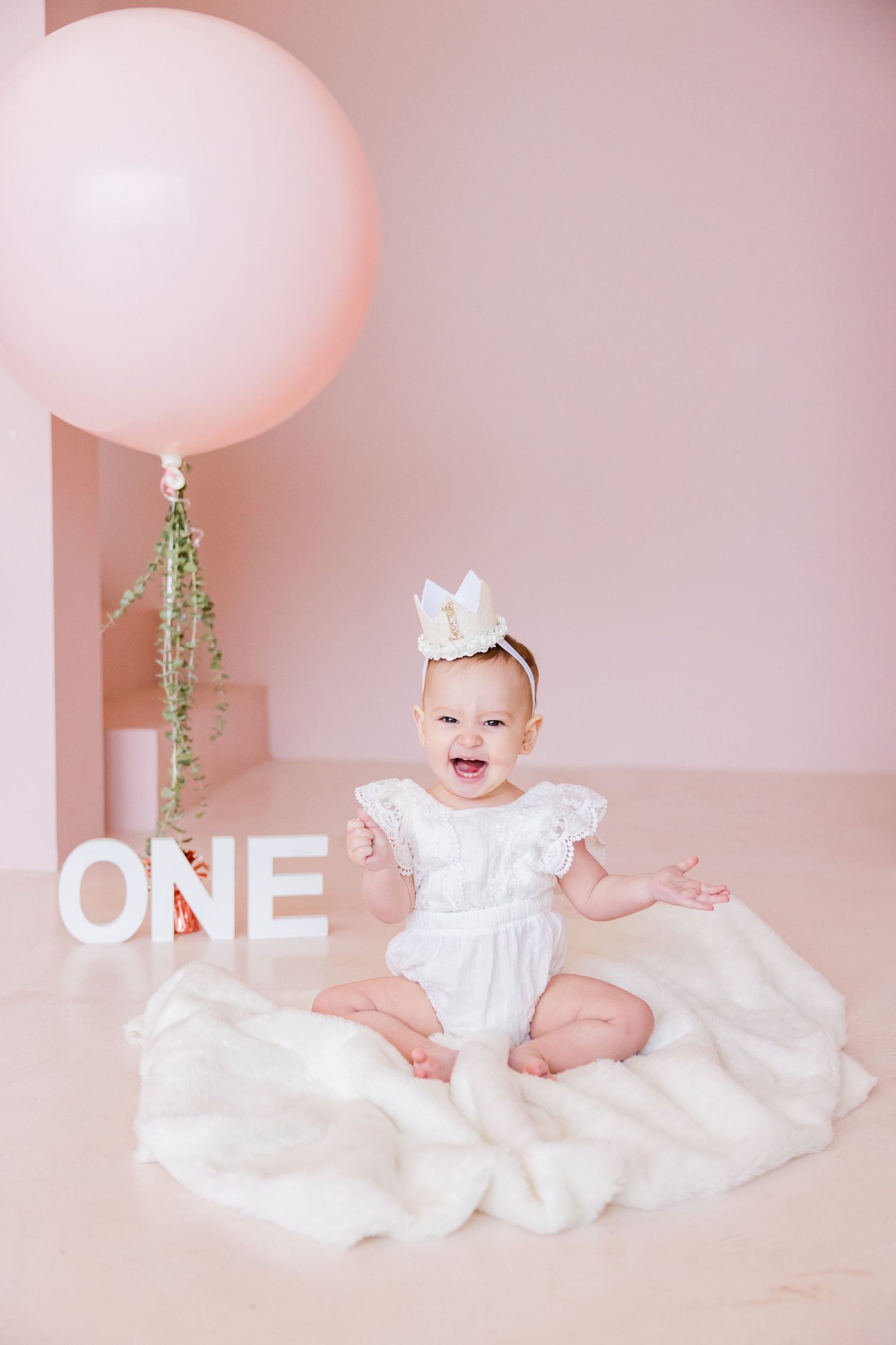 First birthday portrait with large pink balloon, white crown, and white 'ONE' letters on pink backdrop.