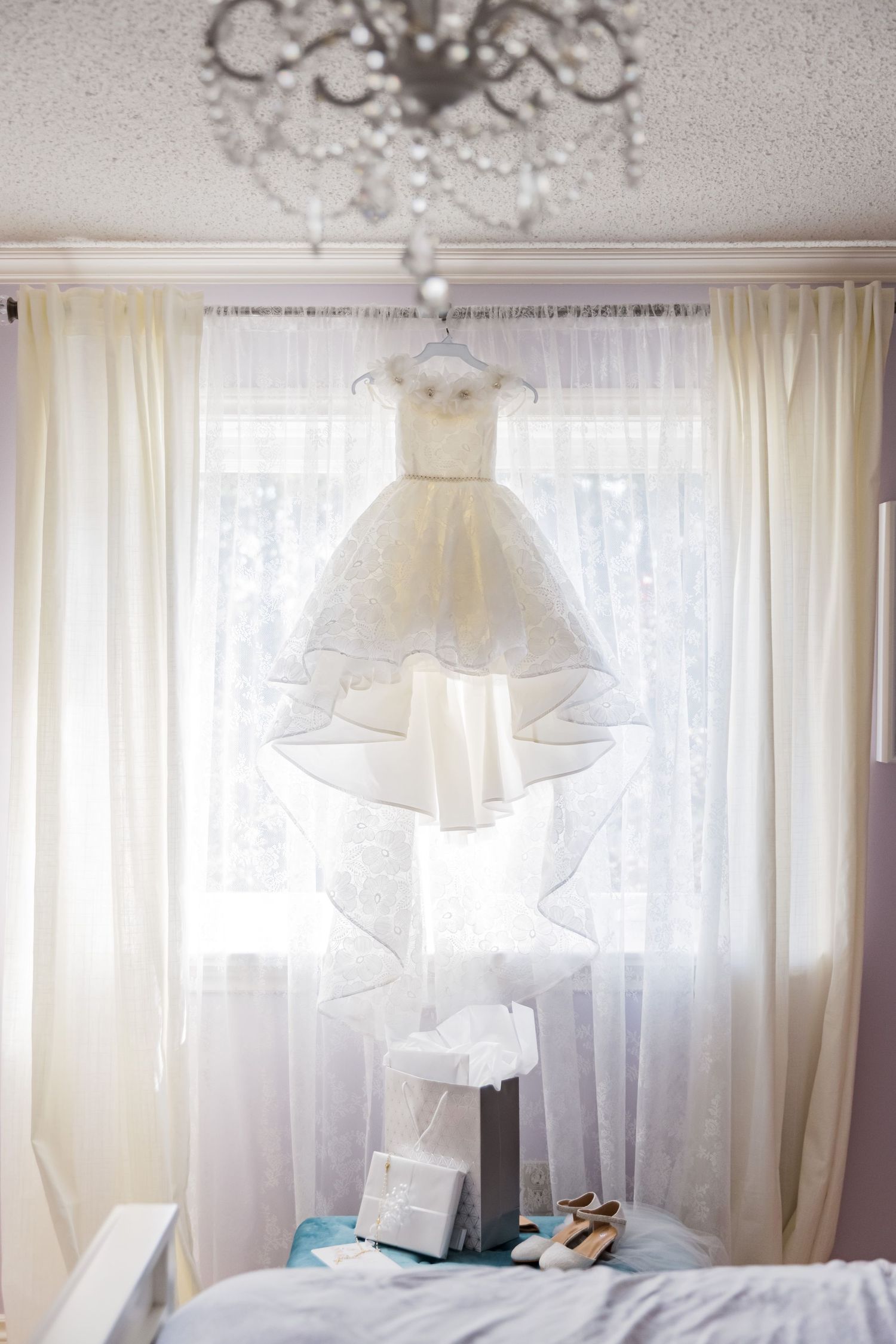 Elegant white wedding dress hanging in front of sheer curtains with crystal chandelier overhead.