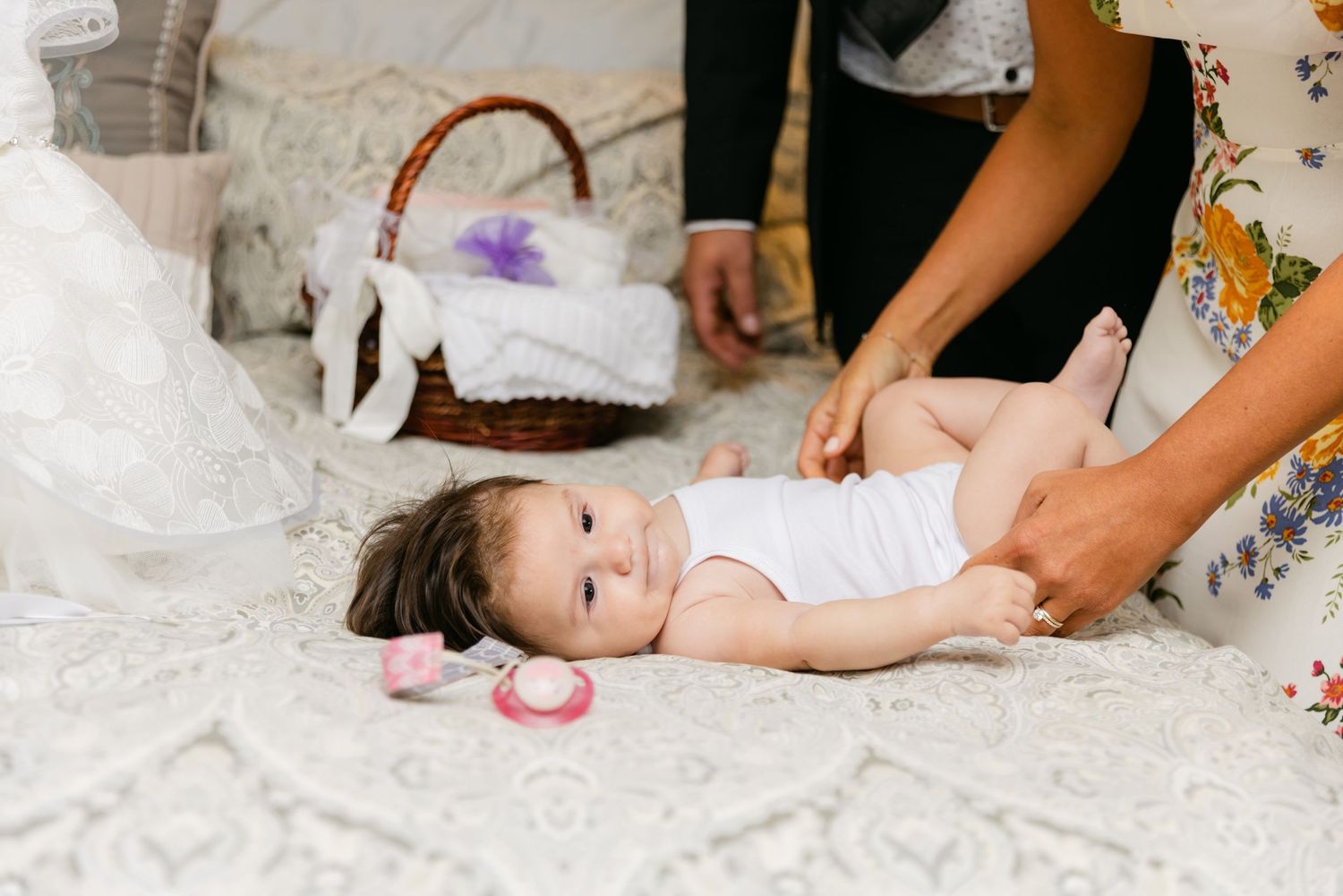 A baby lying on a white bed with hands reaching to adjust their clothing.
